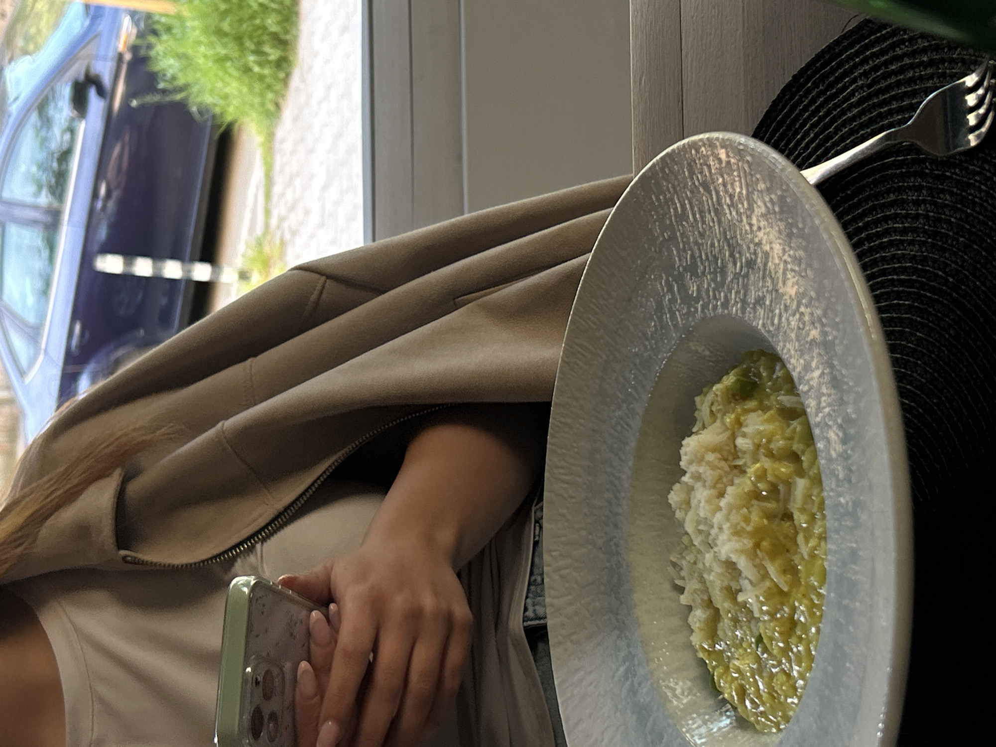 The scene depicts a person sitting at a table in what appears to be a restaurant or café. In the background, visible through a window, is a blue car with greenery. The person is wearing a white top and a tan jacket. Their hand is holding a smartphone with a green case. On the table is a round, light-colored plate containing a dish that appears to be risotto with a light green sauce. A fork rests on a black woven placemat next to the plate. The location is likely in Sofia, Bulgaria.