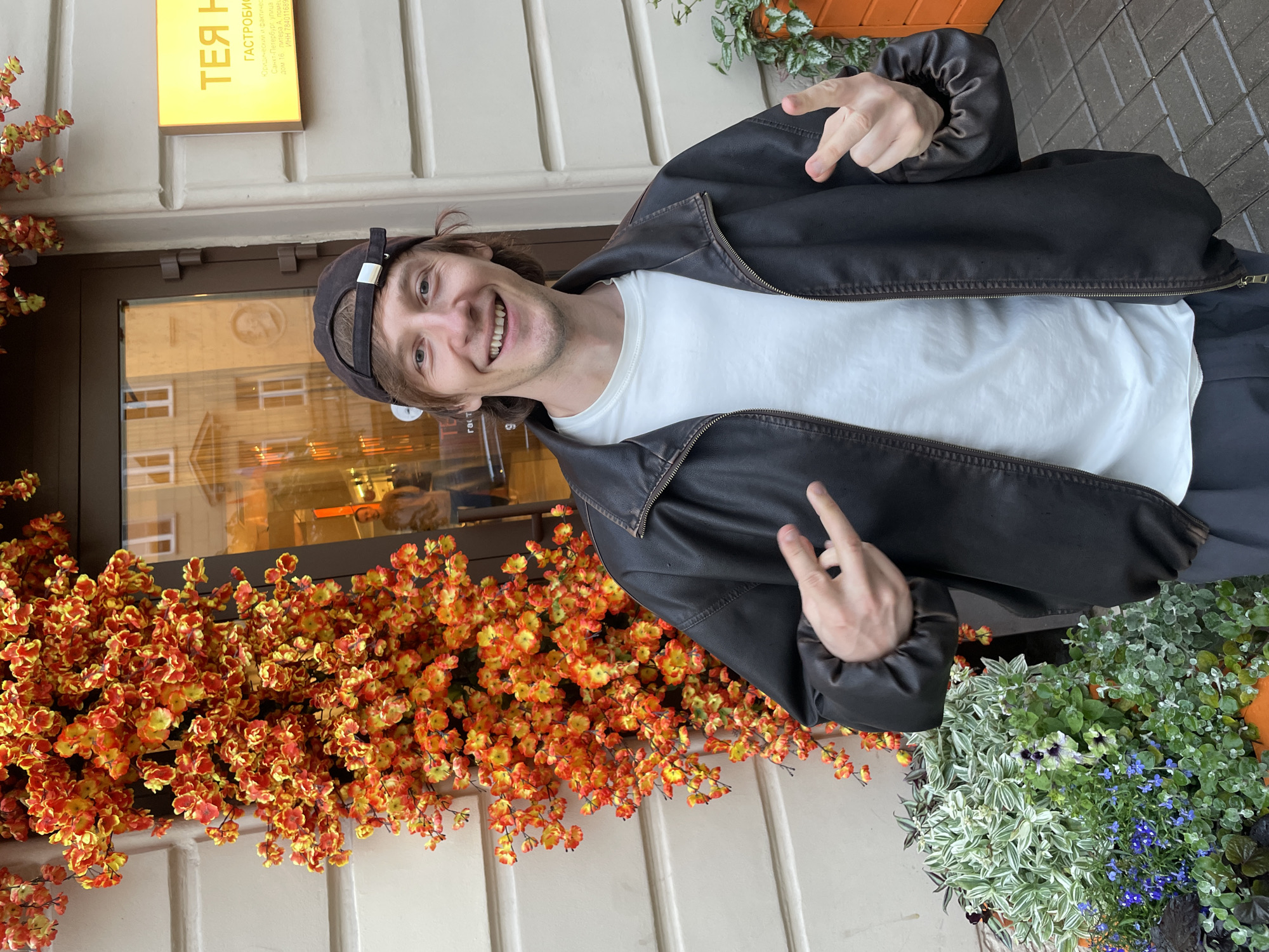 A man wearing a dark brown leather jacket, a white t-shirt, and a baseball cap stands in front of a building with decorative floral arrangements. He is smiling and making a hand gesture with two fingers extended on each hand. Behind him, there is a storefront with a yellow sign and text in Russian. The building's facade is a light color, possibly cream or beige, with architectural details. In the foreground, there are planters filled with greenery and small blue flowers. The ground appears to be paved with cobblestones. The man's reflection is visible in the glass of the storefront. The yellow sign reads "ТЕЯ Н" in large letters, followed by smaller text that includes "ГАСТРОНОМИИ" and an address likely in Saint Petersburg.