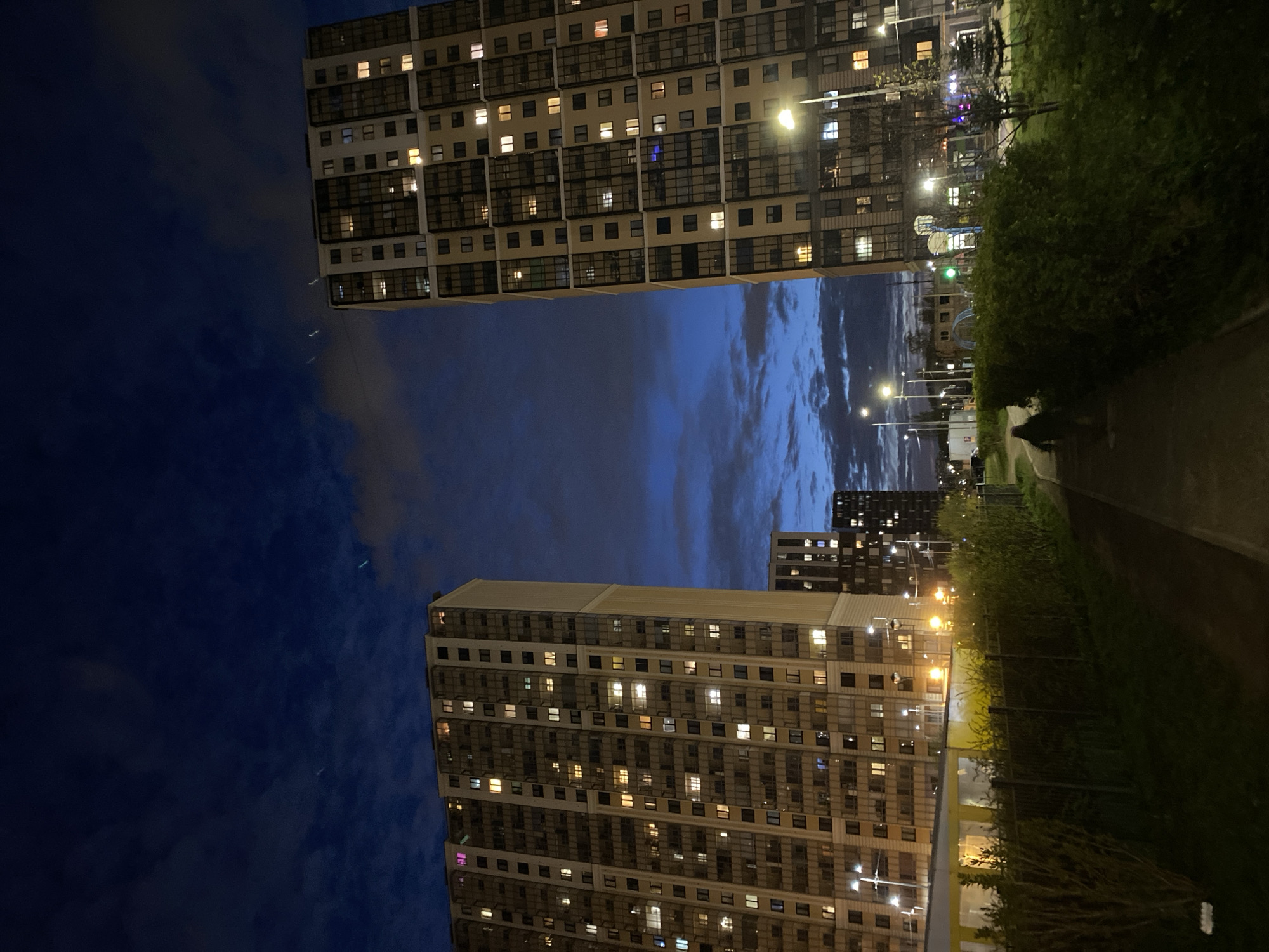 The image depicts a night scene in what appears to be a residential area in Saint Petersburg, Russia, characterized by tall apartment buildings. Two high-rise buildings with many illuminated windows are visible. The buildings are light-colored and have a grid-like pattern of windows, which are lit up, suggesting that the apartments are occupied. Between the buildings, the night sky is visible, showing a gradient of blue and gray clouds.
To the right of the image, there's a walkway or sidewalk lined with greenery, including bushes and trees. Streetlights illuminate the path and the surrounding area. In the background, more buildings are discernible, indicating an urban landscape. A person is walking in the distance on the walkway, casting a faint shadow.