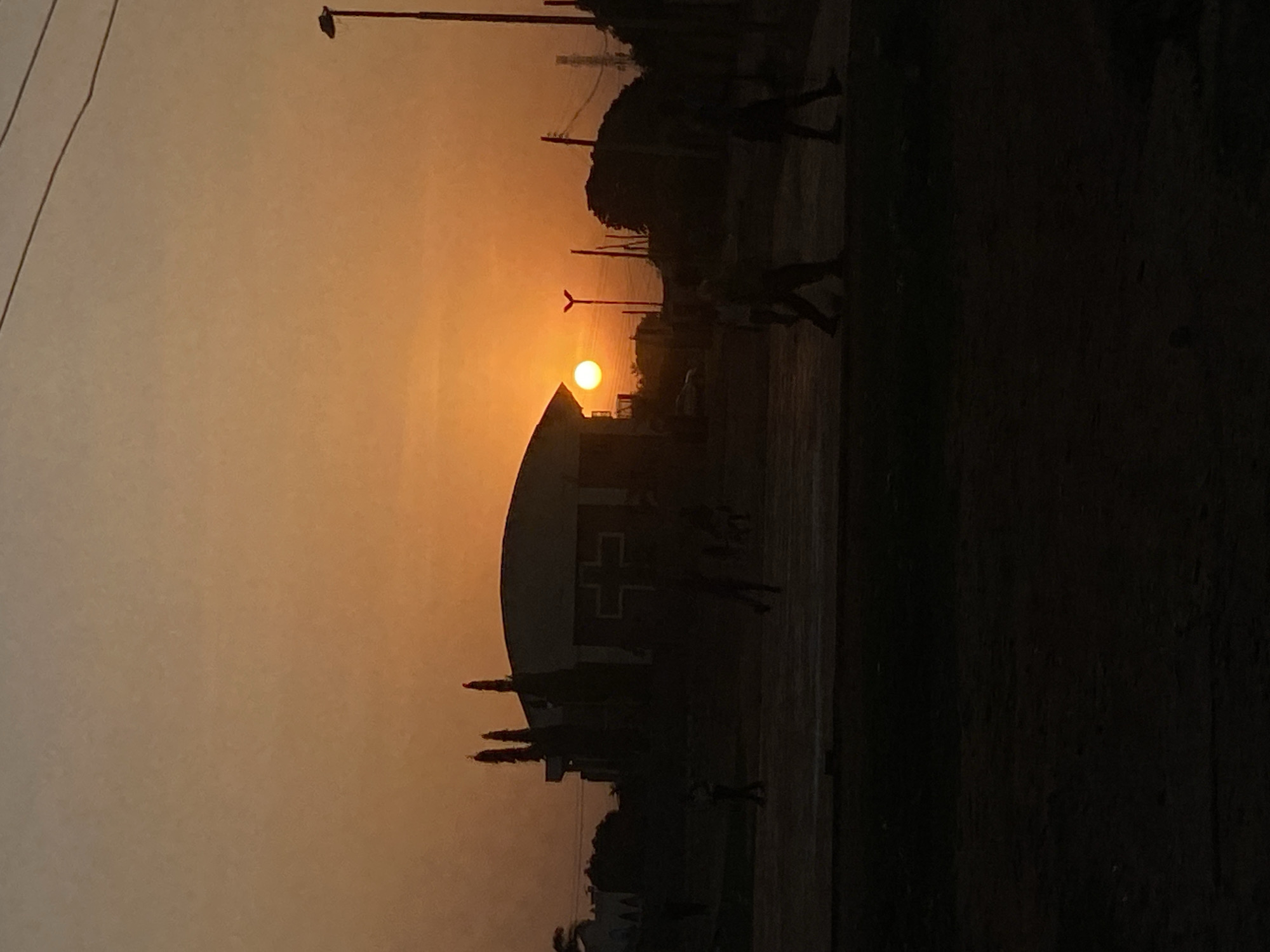 The image captures a sunset scene in Barden Yana, Nigeria. The sun is setting behind a building, casting a silhouette on the structure and surrounding area. The building has a distinctive cross on it, possibly indicating a hospital or church. There are people visible as silhouettes, walking or standing around the building. The environment includes trees, poles, and wires, which are also silhouetted against the sky. The overall scene has a warm, orange hue due to the setting sun. The right side of the image is dark.