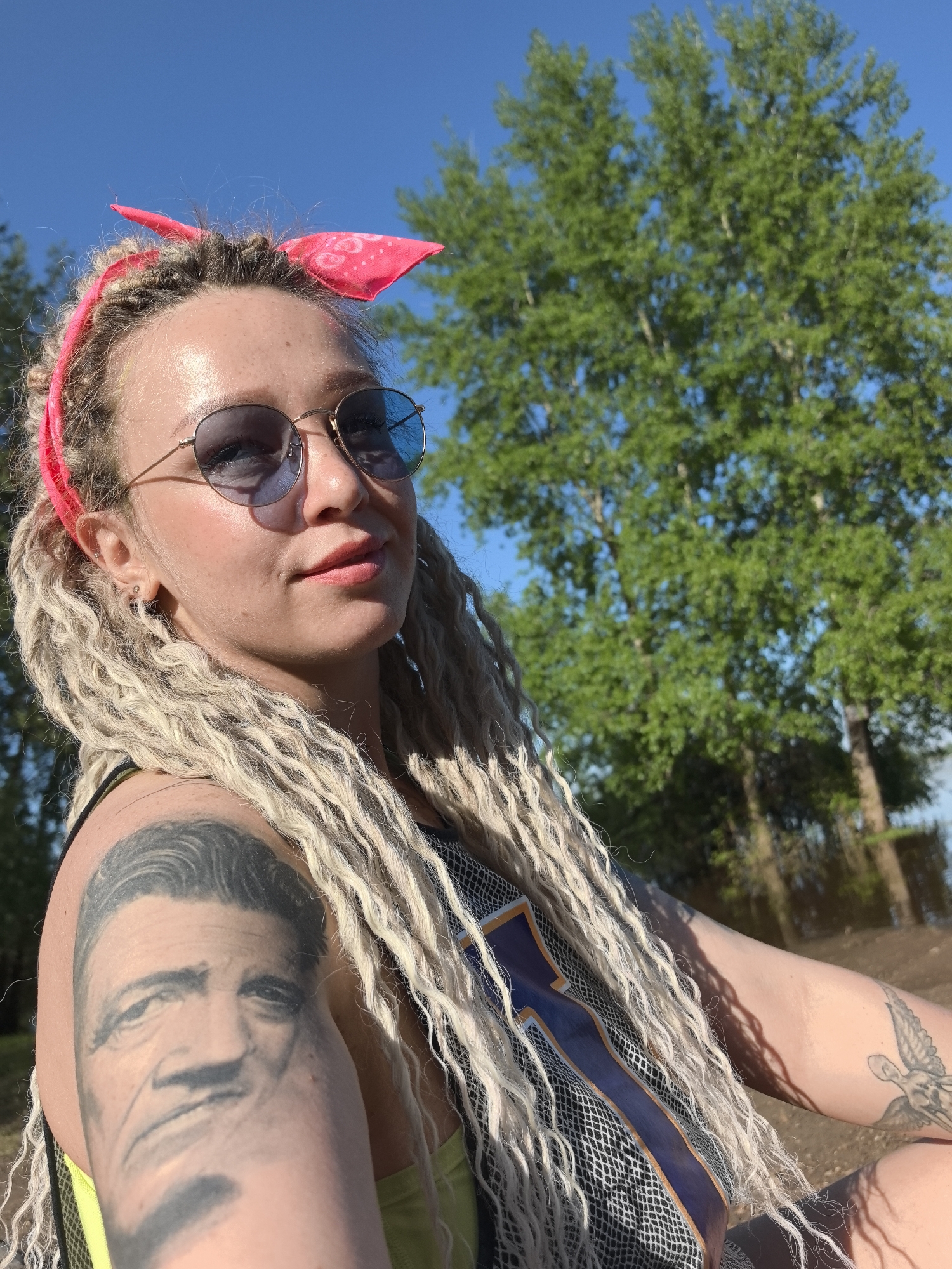 A person with blonde dreadlocks, wearing a pink bandana and round sunglasses, is outdoors with trees in the background under a blue sky. The person has a tattoo of a face on their upper arm and another tattoo of a winged figure on the other arm. The location is implied to be in the city of Noviy, Russia.