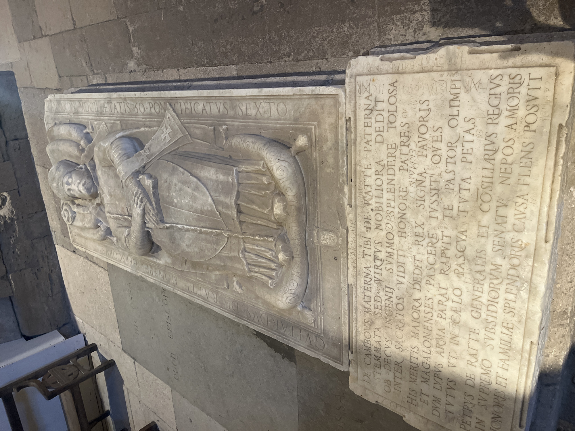This image depicts a stone sarcophagus with an effigy of a man in religious vestments, likely a bishop. The sarcophagus is located within what appears to be a church or cathedral, as indicated by the stone floor and walls. The effigy is detailed, showing the man's face, hands, and clothing, including a mitre on his head and a crozier. Inscriptions are visible on the sarcophagus and on a nearby stone slab. The overall scene is somber and historical, suggesting a place of veneration and remembrance. The sarcophagus is situated on the floor, with a wooden chair visible in the background. The sarcophagus is an important historical artifact, likely dating back several centuries, and is located in Villeneuve-lès-Maguelone, France.