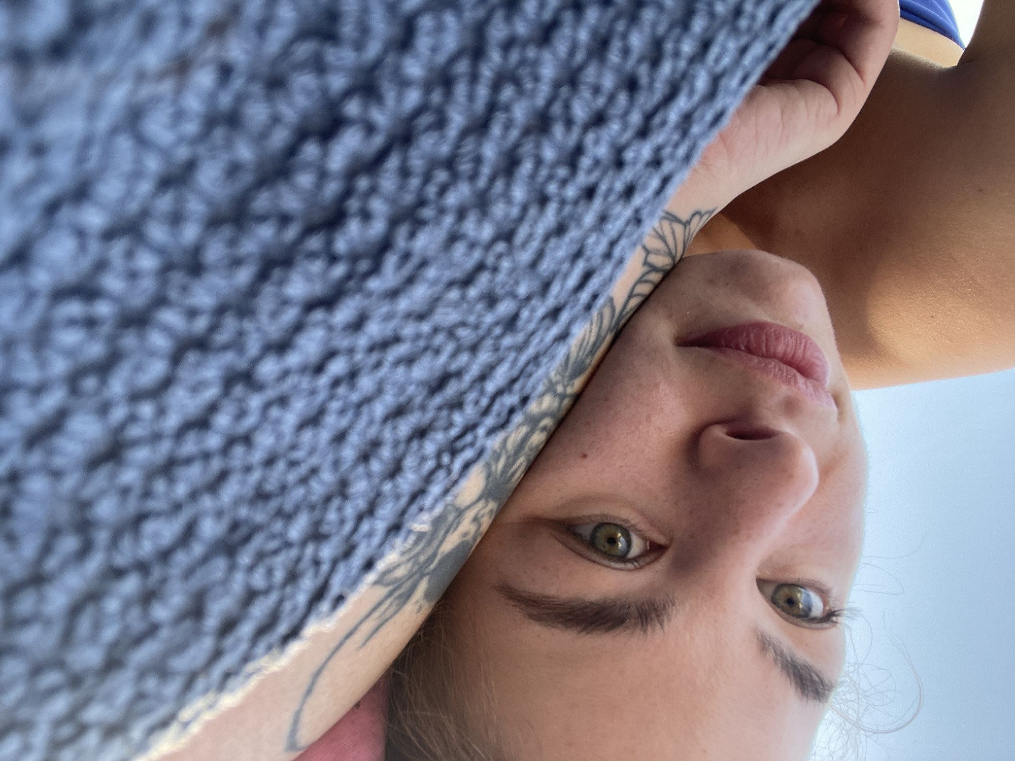 The image is a close-up, rotated selfie featuring a person with fair skin, green/hazel eyes, and dark eyebrows. The person's face is oriented upside down and diagonally, with their head tilted to the right. A prominent blue-grey knitted or crocheted fabric, likely a blanket or towel, covers a significant portion of the upper left side of the frame, partially obscuring the person's forehead and hair.
Below the fabric, a tattoo featuring floral or leaf-like patterns is visible on the person's left arm or shoulder. Their left hand is also partially seen near their face. The person's lips are slightly parted, hinting at a subtle smile, and their eyes are open, looking directly at the camera with a neutral expression. A small section of a blue garment, possibly a swimsuit, is visible in the upper right corner of the image. The background is a bright, clear light blue, strongly suggesting an open sky.
The scene conveys a moment of relaxation outdoors during daytime, likely in a sunny environment. Given the location of Rafailovići, Montenegro, the setting is presumably a beach or coastal area under a clear sky. There is no visible text in the image.