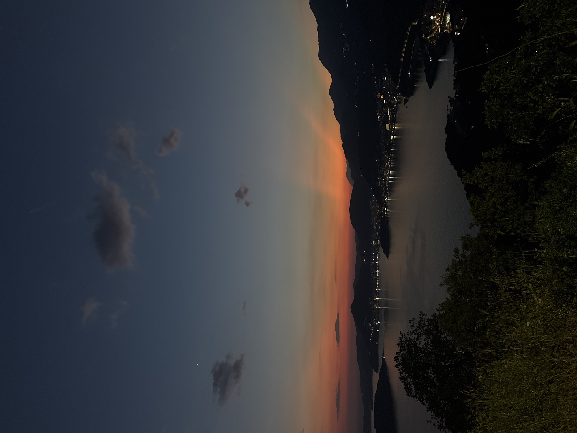 The image shows a twilight scene of Kamenovo, Montenegro, from an elevated perspective. In the foreground, dark vegetation is visible on the right side. A body of water, possibly the Adriatic Sea, stretches into the distance, reflecting the lights of the city. On the opposite side, there is a mountain range silhouette. The sky transitions from dark blue on the left to orange and pink hues near the horizon. A few scattered clouds are in the sky.