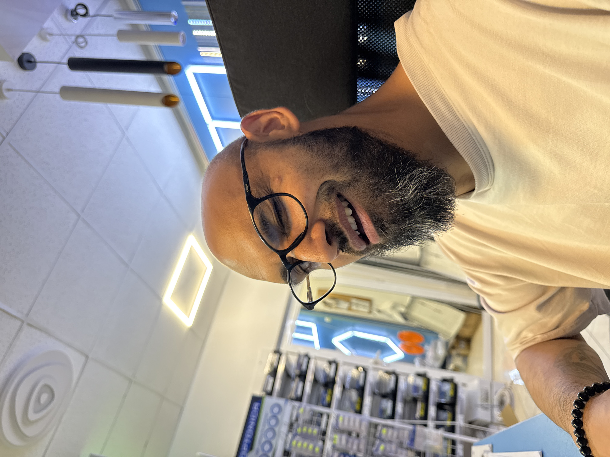 The image depicts an adult male captured mid-laugh or smile, seated in what appears to be a lighting showroom or office of Optonior Ltd. in Plovdiv, Bulgaria. The man, with a bald head and dark skin, wears dark-rimmed "CARRERA" eyeglasses and a light, possibly off-white or cream-colored, short-sleeved crew-neck t-shirt. He has a full, dark beard with significant graying, particularly visible on the chin and sides. His eyes are squinted shut, and his mouth is wide open in an expression of mirth, revealing his tongue. A black beaded bracelet is visible on his left wrist. He is positioned in front of a dark, possibly mesh-backed chair.
The background, predominantly in shades of white, blue, and light gray, indicates an indoor commercial setting. To the left, metal display shelving units hold numerous packaged items, likely lighting components or electronics, many in clear plastic with yellow labels. A partial blue and white sign bearing the name "OPTONIOR" is visible above these shelves. The ceiling is a white acoustic panel grid system with various lighting fixtures: a prominent, bright rectangular LED panel, a spiral-patterned recessed vent, and several modern cylindrical pendant lights hanging down. Two pendant lights are light-colored (off-white/cream), and one is black, all exhibiting a warm orange glow from their open bottoms. A light blue wall or partition runs horizontally across the mid-background, featuring bright blue neon light installations, including one resembling a stylized letter, and fainter orange neon elements further back.