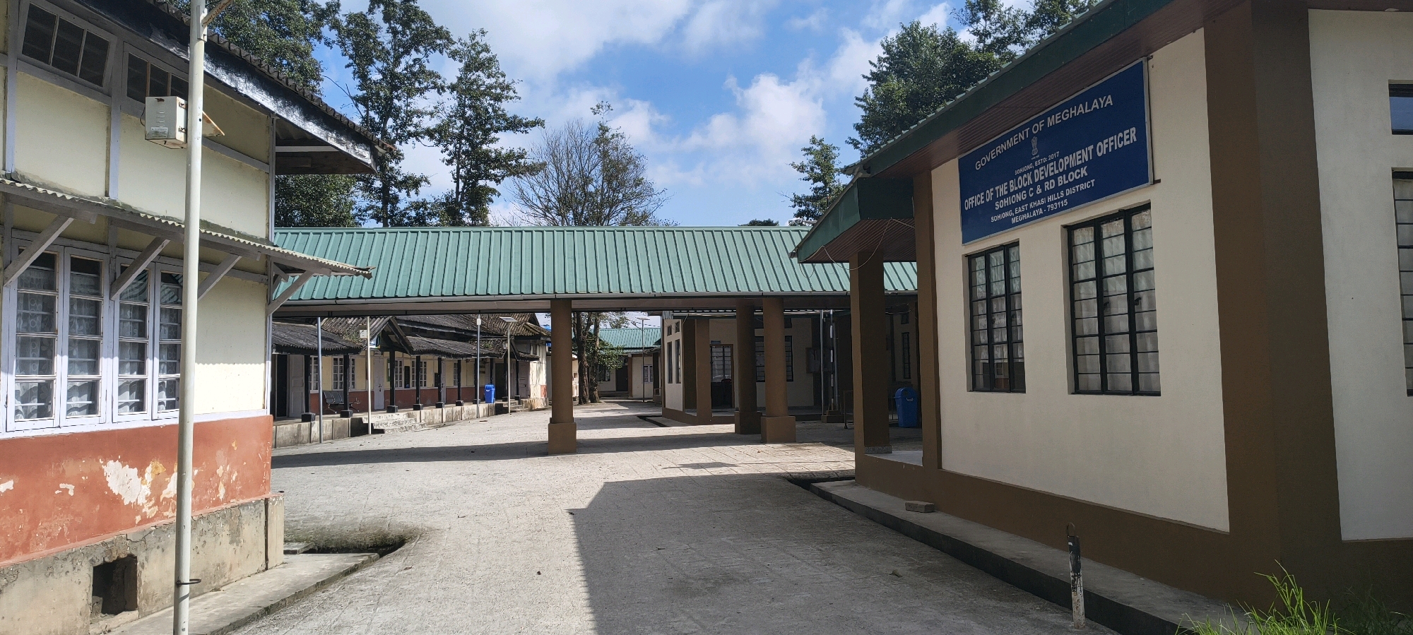 The image shows a view of the Office of the Block Development Officer in Sohiong, East Khasi Hills District, Meghalaya, India. The building is a single-story structure with a beige exterior and brown trim around the windows and base. The blue sign on the building reads "GOVERNMENT OF MEGHALAYA OFFICE OF THE BLOCK DEVELOPMENT OFFICER SOHIONG, ESTD: 2017 SOHIONG C & RD BLOCK SOHIONG, EAST KHASI HILLS DISTRICT MEGHALAYA - 793115". There are large, black-gridded windows on the building. A covered walkway with a green corrugated metal roof connects this building to other structures on the property. In the foreground, a paved courtyard is visible, with some grass growing at the edges. Several trees are present in the background, with a blue sky and scattered clouds above. No people are visible in the image.