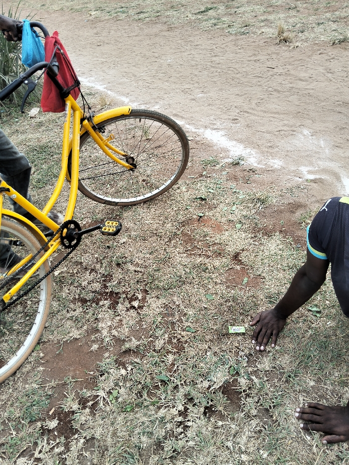 The image depicts an outdoor scene in Waa Ngombeni ward, Kenya. The ground surface consists of reddish-brown soil with sparse patches of dry, light green and brown grass. A distinct white line is visible on the dirt ground in the upper right portion of the frame, extending horizontally.
A bright yellow bicycle is partially present on the left side, angled with its front wheel facing the upper right. A blue plastic bag and a red fabric bag are suspended from the black handlebars. The bicycle has yellow mudguards, a visible chain, and a black pedal with yellow accents. The lower legs of an individual, wearing dark trousers, are visible next to the bicycle's frame on the far left.
On the right side of the frame, another individual is partially visible, appearing to be kneeling or crouching. This person wears a dark blue shirt with light blue and yellow trim on the right sleeve. Their dark-skinned hands are placed palms down on the ground. Directly in front of their hands, a small, rectangular, green object rests on the soil amidst the grass.
