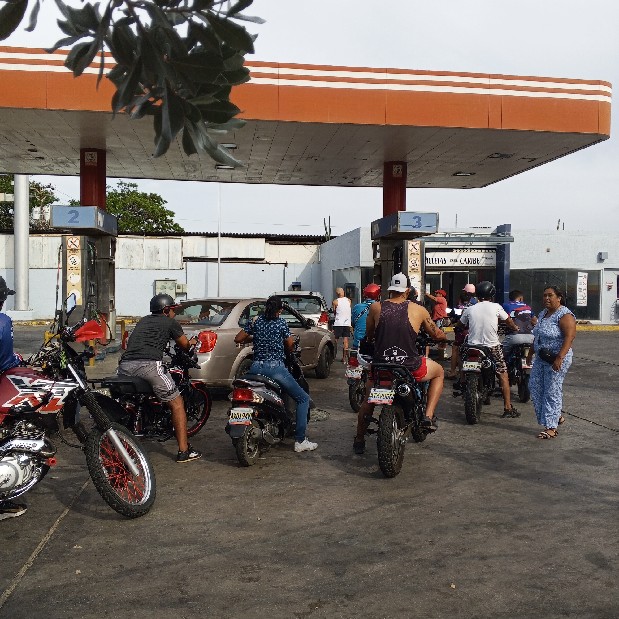 The image captures a scene at a gas station in Unknown, Unknown. Several motorcycles and a car are lined up at the pumps under the station's orange and white canopy. A woman in a blue dress stands near the line of motorcycles, observing the scene. Several people on motorcycles are waiting in line. The license plates of a few vehicles are visible. In the background, a building with "BICICLETAS DEL CARIBE" signage can be seen. The general atmosphere appears to be one of waiting and activity.