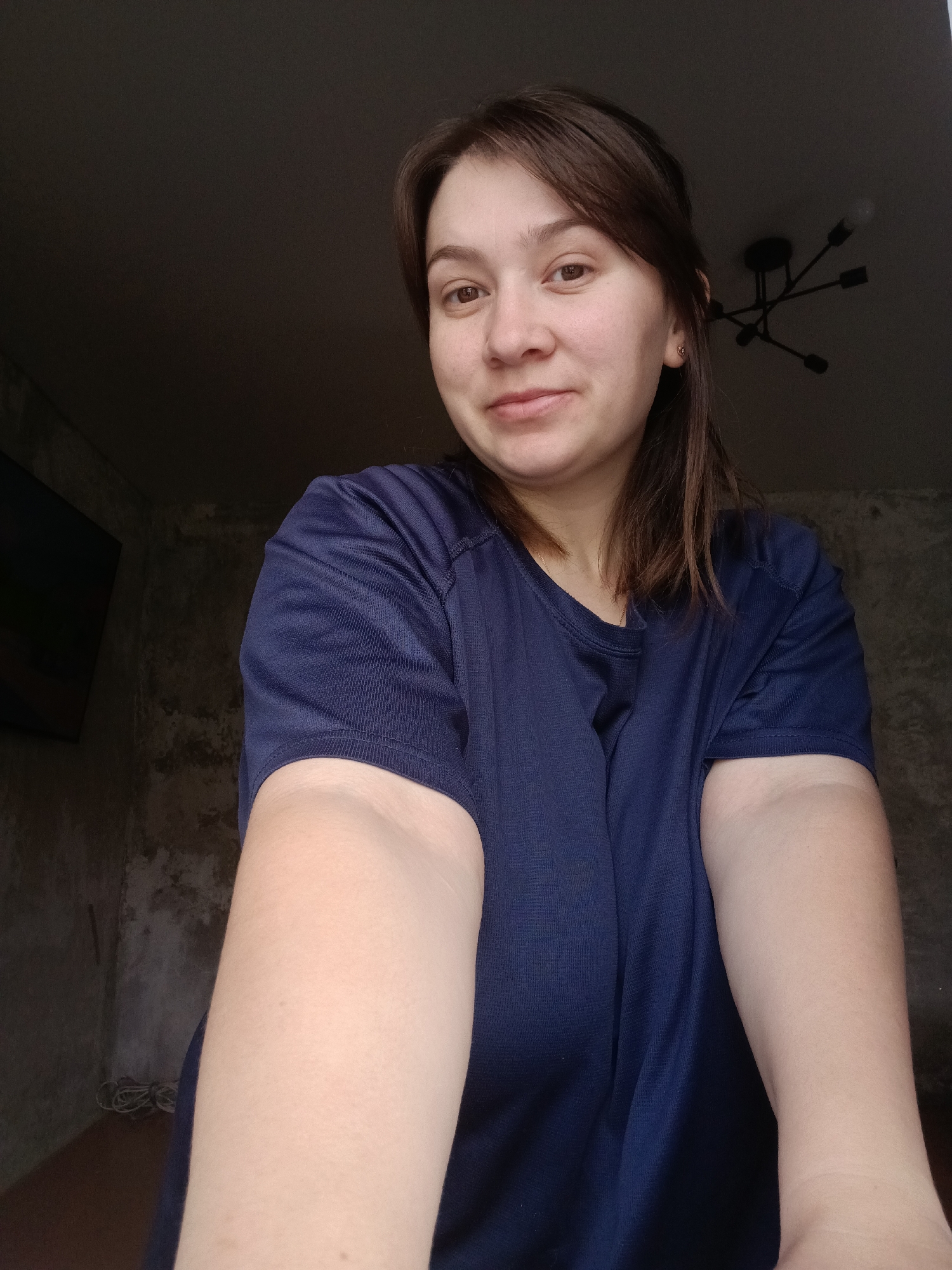 A woman with light brown hair and brown eyes is looking at the camera. She is wearing a dark blue t-shirt. She is indoors, in a room with a dark, textured wall and a dark ceiling. A modern black chandelier is visible on the ceiling. There is a television screen on the left side of the frame. The lighting in the room is dim. The image appears to be a selfie.