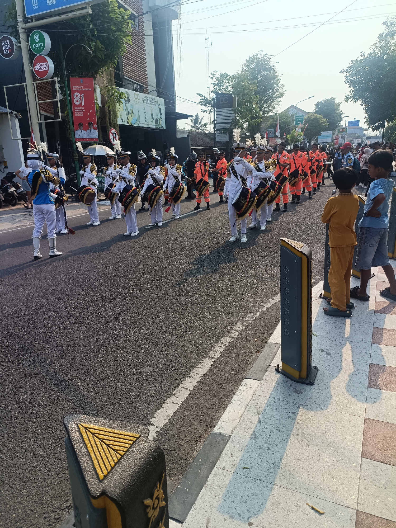 This image captures a marching band parade on a street in Sleman, Indonesia. The band members, dressed in white and blue uniforms with feathered hats, are playing drums. In the background, several buildings with signage are visible, including "SAY BRE AD," "POINT COFFEE," and "Yummy Choice." A large red banner with "DIRGAHAYU REPUBLIK INDONESIA 81" is displayed on one of the buildings, indicating a celebration of Indonesia's 81st Independence Day. Spectators, including children, are gathered on the sidewalk, observing the parade. The sun is shining, casting shadows on the street and sidewalk. The scene appears to be a festive public event taking place in a commercial area of Sleman.