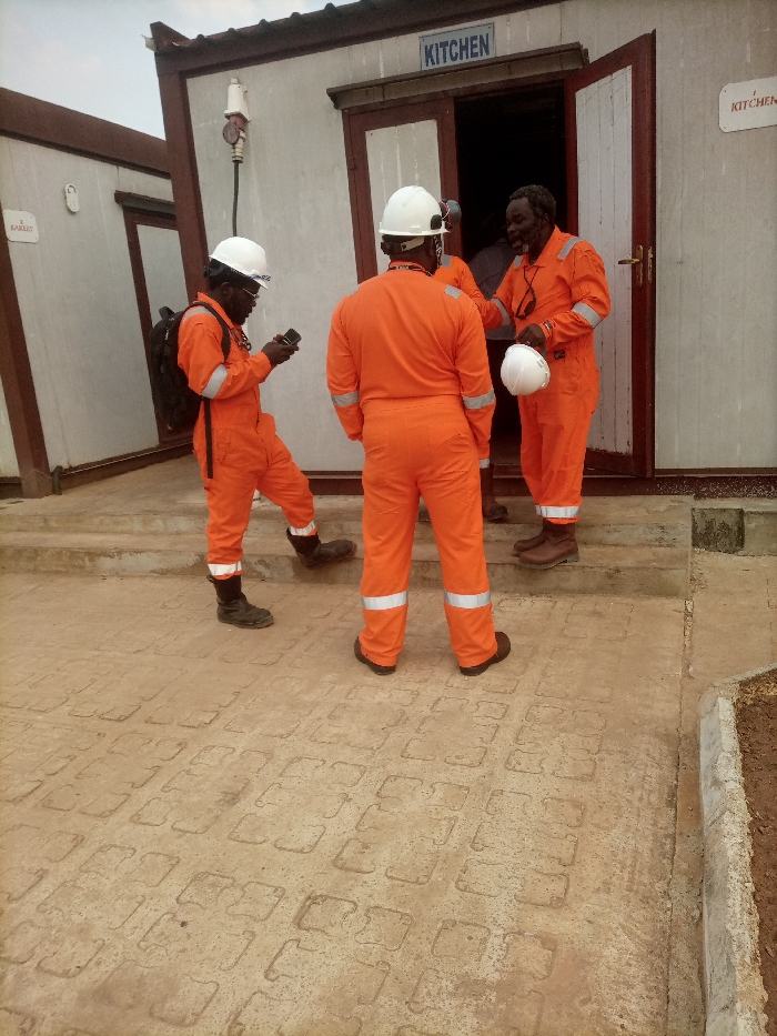 The scene, located in Opete, Nigeria, depicts three individuals, identified as black males, wearing orange work coveralls with reflective stripes and white hard hats, standing on a paved area outside two modular buildings under an overcast sky.
The primary building on the right is light-colored with brown trim and has a partially open brown door. A blue sign with white text above this door reads "KITCHEN." A smaller, identical "KITCHEN" sign is present further right on the building's facade. An electrical outlet with a red plug is mounted on the exterior wall to the left of the door.
On the left side of the frame, one male subject wears a black backpack and black safety boots. He holds and views a black smartphone with both hands, with his right foot raised onto a concrete step leading to the "KITCHEN" building. A second male subject stands centrally with his back to the camera, facing the "KITCHEN" doorway. The third male subject, an older individual with a beard, stands at the threshold of the "KITCHEN" doorway, holding a white hard hat in his left hand, with dark glasses hanging from his neck. He is facing the central figure, smiling, indicating a social interaction.
The ground is paved with interlocking light-colored concrete blocks, showing signs of wear and dirt. A concrete step provides access to the "KITCHEN" building entrance. A second modular building, partially visible on the far left, features a sign with text that partially reads "BAKERY." A low concrete retaining wall bordering disturbed earth is visible on the far right.