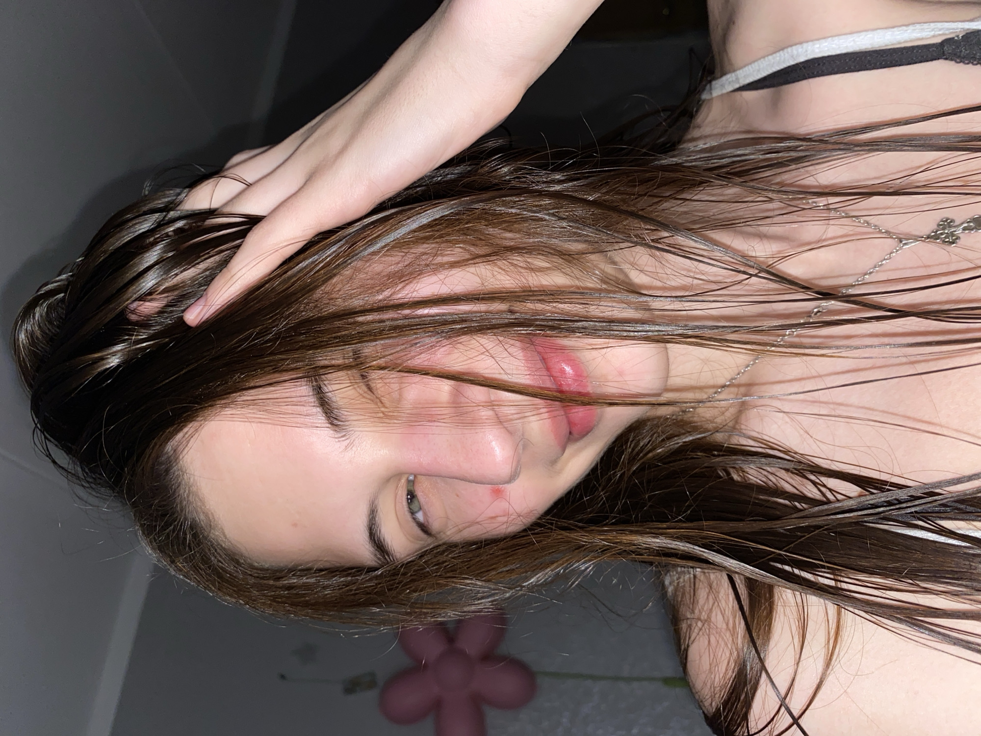 A young woman with wet, brown hair is lying down, possibly in an indoor setting. She has fair skin, a small mark near her nose, and is wearing a necklace. Her hand is touching her hair. There is a small pink, flower-shaped object visible in the background. The image is taken from a slightly downward angle and is rotated. The location cannot be determined with the available information, and the city of Votkinsk, Russia, cannot be confirmed.