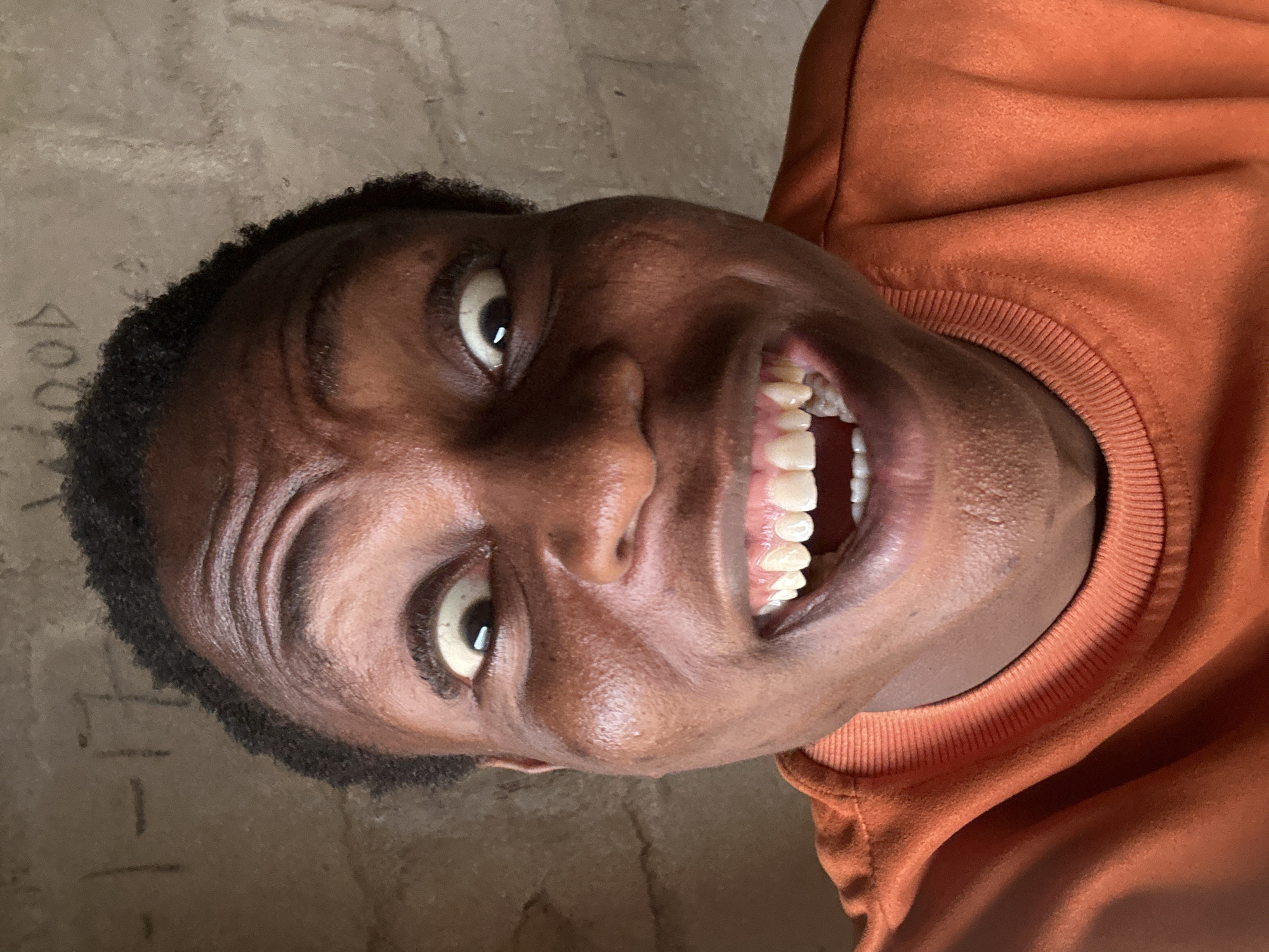 A dark-skinned person with short black hair, wearing an orange sweatshirt, is smiling broadly. The background is a textured beige wall with some indistinct markings. The location appears to be indoors, possibly in the city of Unknown, Unknown.