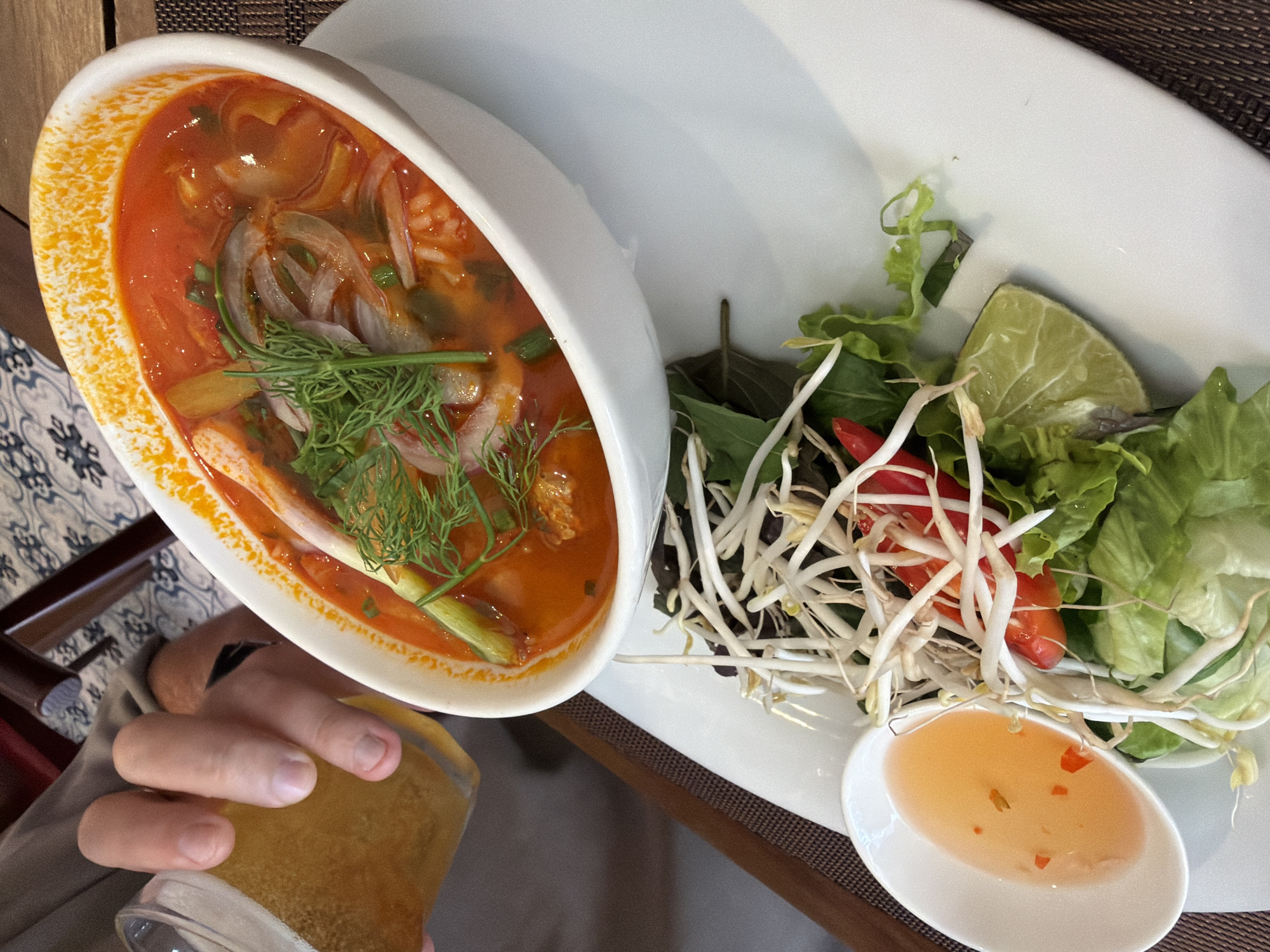 A close-up overhead shot shows a person's hand holding a glass of golden beer, positioned above a white bowl of pho soup. The soup is a vibrant red broth filled with noodles, sliced onions, green herbs, and what appear to be pieces of meat. Beside the soup bowl, on a white plate, is a garnish of fresh lettuce, bean sprouts, sliced red chili peppers, a lime wedge, and a small white dish containing a light orange dipping sauce. The setting appears to be a restaurant in Nha Trang Ward, Vietnam, suggested by the traditional patterned tablecloth and wooden furniture visible in the background.