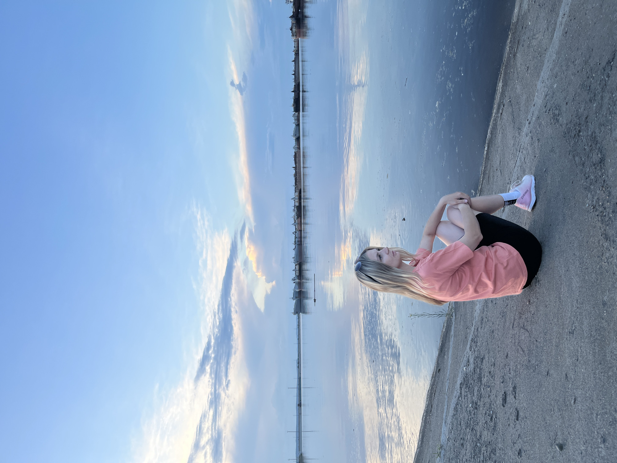 A young woman with long blonde hair sits on a concrete embankment, facing right, with her knees drawn up to her chest. She is wearing a coral-colored t-shirt and black shorts, and pink sneakers. The embankment slopes down to a wide body of water, likely the Kama River, given the location of Chaikovsky, Russia, in the background. Several barges and ships are docked in the distance, lined up along the horizon. The sky above is a pale blue with wispy clouds, some of which are illuminated by the setting or rising sun, casting reflections on the calm water. The overall atmosphere is peaceful and serene.