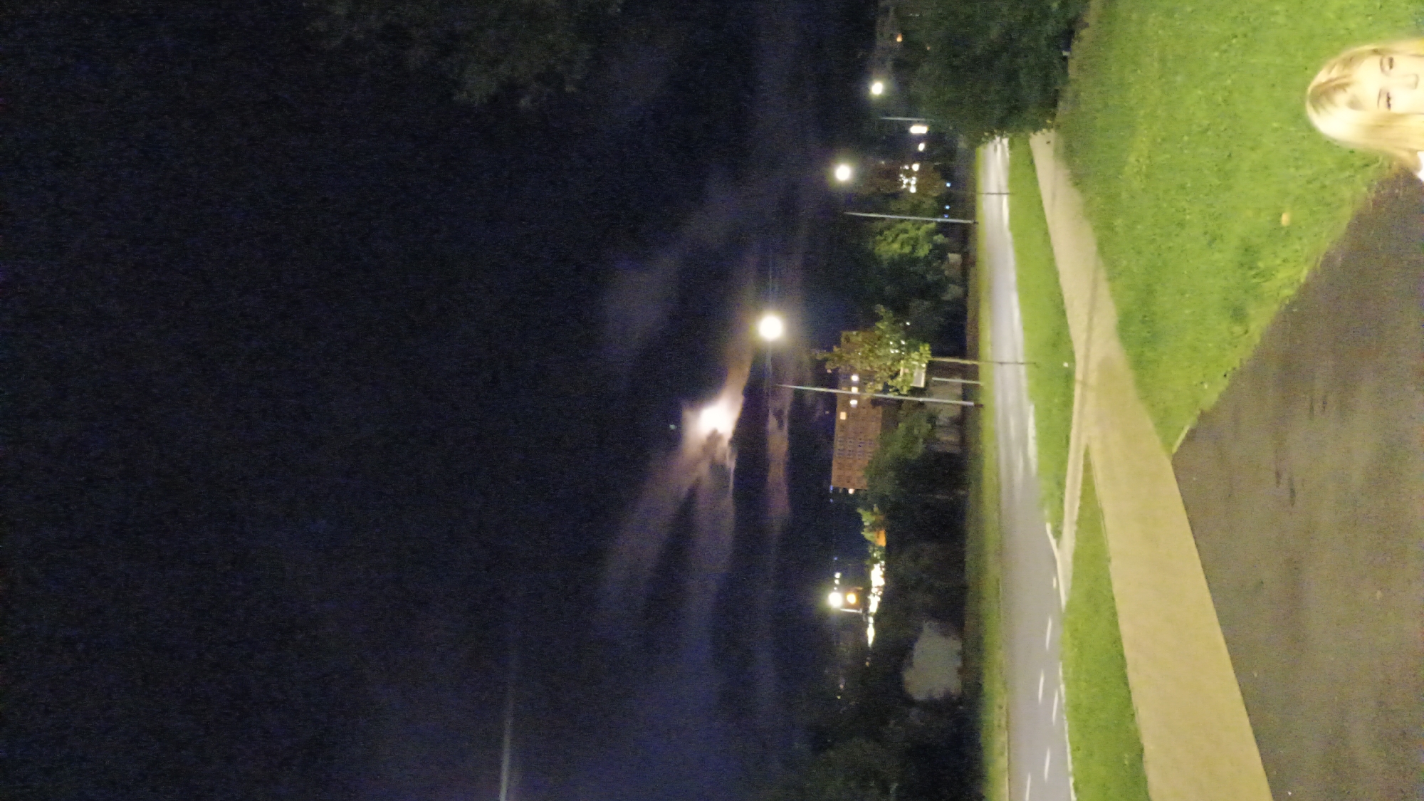 The image captures a nighttime scene on a street in Sokolov, Czechia. The sky is dark with some wispy clouds illuminated by a bright moon, which is partially obscured by the clouds, creating a luminous effect. A street lamp is visible to the right, casting a bright light. In the background, there are several buildings with lights on, suggesting an urban environment. A street with white lane markings runs horizontally across the mid-ground, bordered by grassy areas and a paved walkway in the foreground. A young woman with blonde hair is partially visible in the bottom right corner of the frame, looking towards the camera. The overall impression is one of a quiet, urban night.