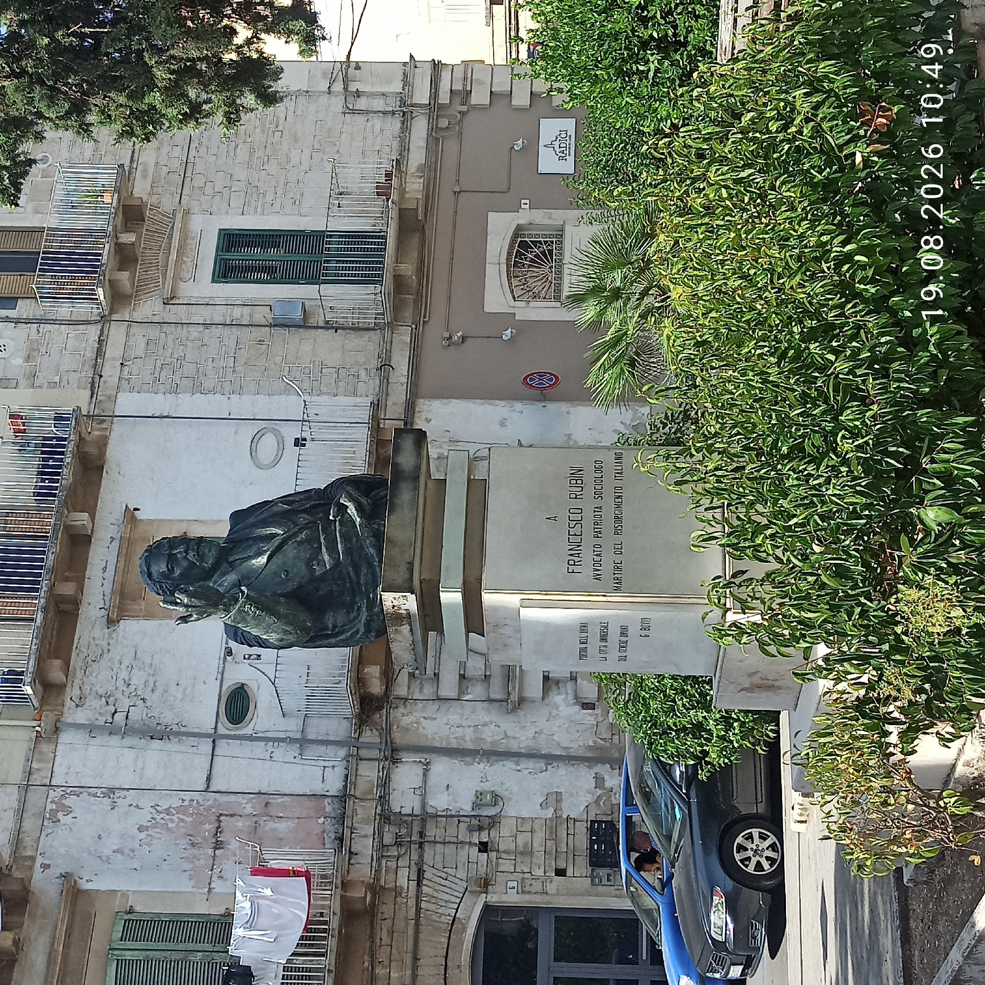 The image shows a bronze statue of Francesco Rubini, a lawyer, patriot, sociologist, and martyr of the Italian Risorgimento, located in front of a building in Ruvo di Puglia, Italy. The statue depicts Rubini standing and raising his right hand in a gesturing or saluting pose. He is wearing a suit and tie. The statue is mounted on a large, multi-tiered stone pedestal which has an inscription on its front face. The building behind the statue is a multi-story structure with balconies and windows, some of which have green shutters. Laundry is hanging from one of the balconies. In the foreground, there is dense green foliage. Two cars are visible in the lower left of the image, and a street sign is partially visible on the right. The timestamp "19.08.2026 10:49" is present in the bottom right corner.