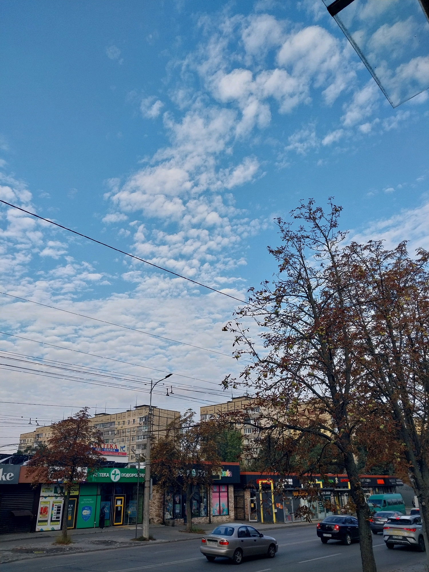 The image depicts a street scene in Dnipro, Ukraine. A blue sky with scattered white clouds serves as the backdrop. In the foreground, trees with autumn foliage are visible. A street runs horizontally across the image, with cars driving on it. On the right side of the street, a row of storefronts is visible, including a pharmacy with a green sign and a white cross logo. Behind the storefronts, apartment buildings are seen. Power lines are strung across the sky. The overall impression is a typical urban streetscape on a cloudy autumn day.