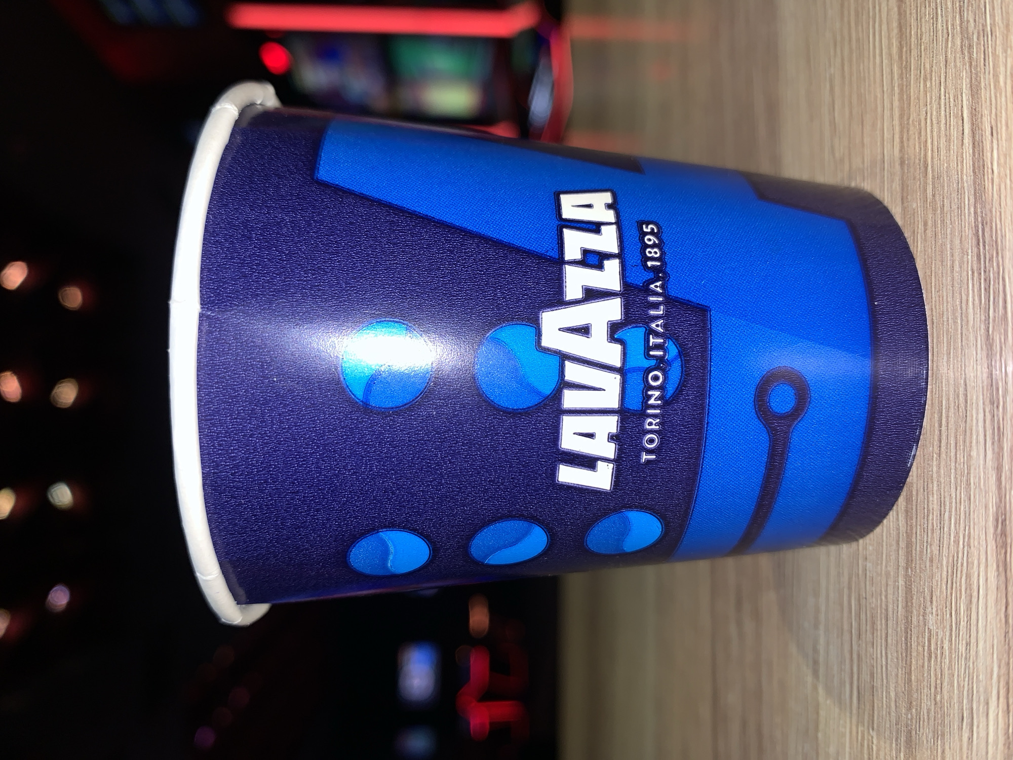 A close-up, eye-level, medium shot captures a disposable paper cup with a dark blue textured exterior adorned with a bright blue geometric design and white "LAVAZZA" text, with "TORINO. ITALIA. 1895" below. The cup is positioned on a light-brown wooden surface with a visible grain. The background is dark and blurred, featuring colorful, out-of-focus lights, possibly from an arcade or entertainment venue. There are no people visible in the image, and no specific location in Burgas, Bulgaria, can be identified.
