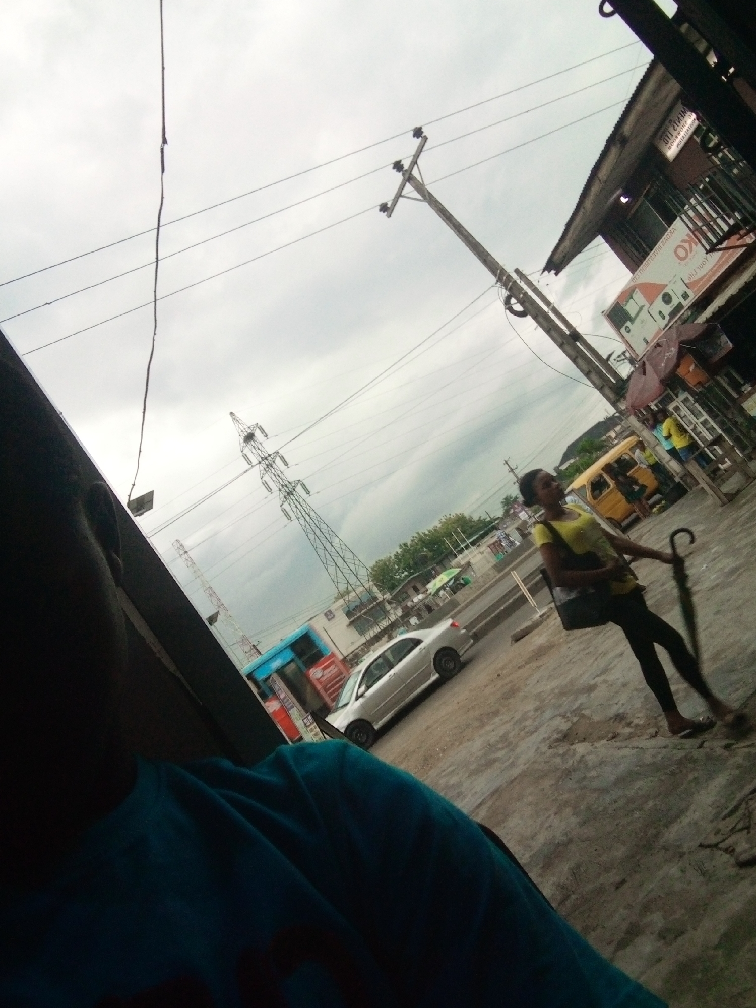 The image depicts a street scene in Shangisha, Nigeria, under an overcast sky. In the foreground, a person wearing a bright blue sweatshirt is partially visible, with their face obscured. A young woman in a yellow t-shirt and black leggings walks away from the camera, holding a dark umbrella. She appears to be carrying a dark bag over her shoulder. Behind her, a yellow van is parked, and a blue and red bus is visible further down the street. Various buildings, some with corrugated metal roofs, line the street. Electrical wires and utility poles are prominent features of the urban landscape, along with a large lattice metal tower in the distance. The ground is paved with concrete and dirt.