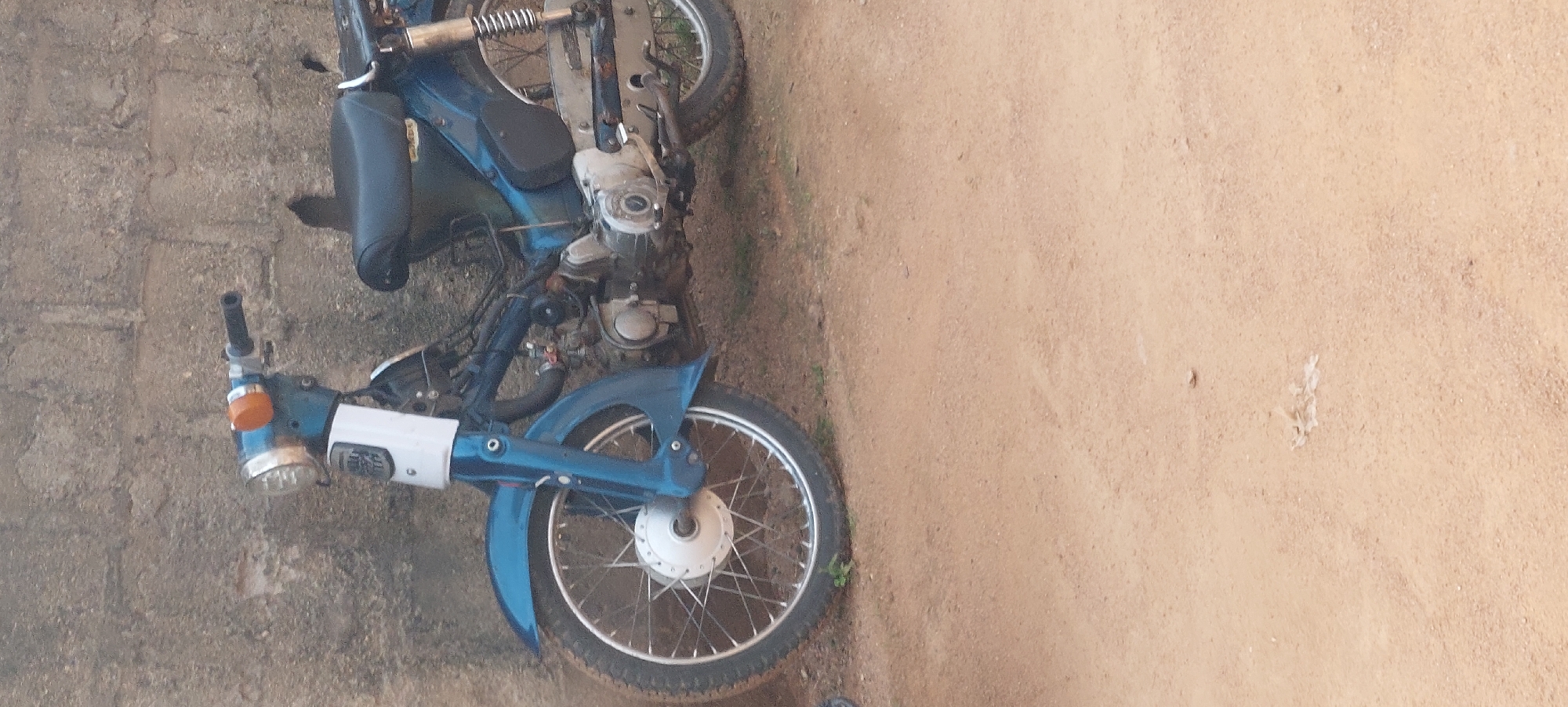 A blue and white Honda C50 motorcycle is parked on a dirt road in front of a textured concrete wall. The motorcycle is facing left, with its front wheel and part of the engine visible. The seat is black, and there is a silver rack on the back. The handlebars and front headlight assembly are also visible. The wall appears to be made of roughly mortared bricks and shows signs of wear. The ground is a sandy, reddish-brown dirt. There are no people or animals visible in the image. The scene suggests an outdoor, possibly rural or semi-urban setting in Jalingo, Nigeria.