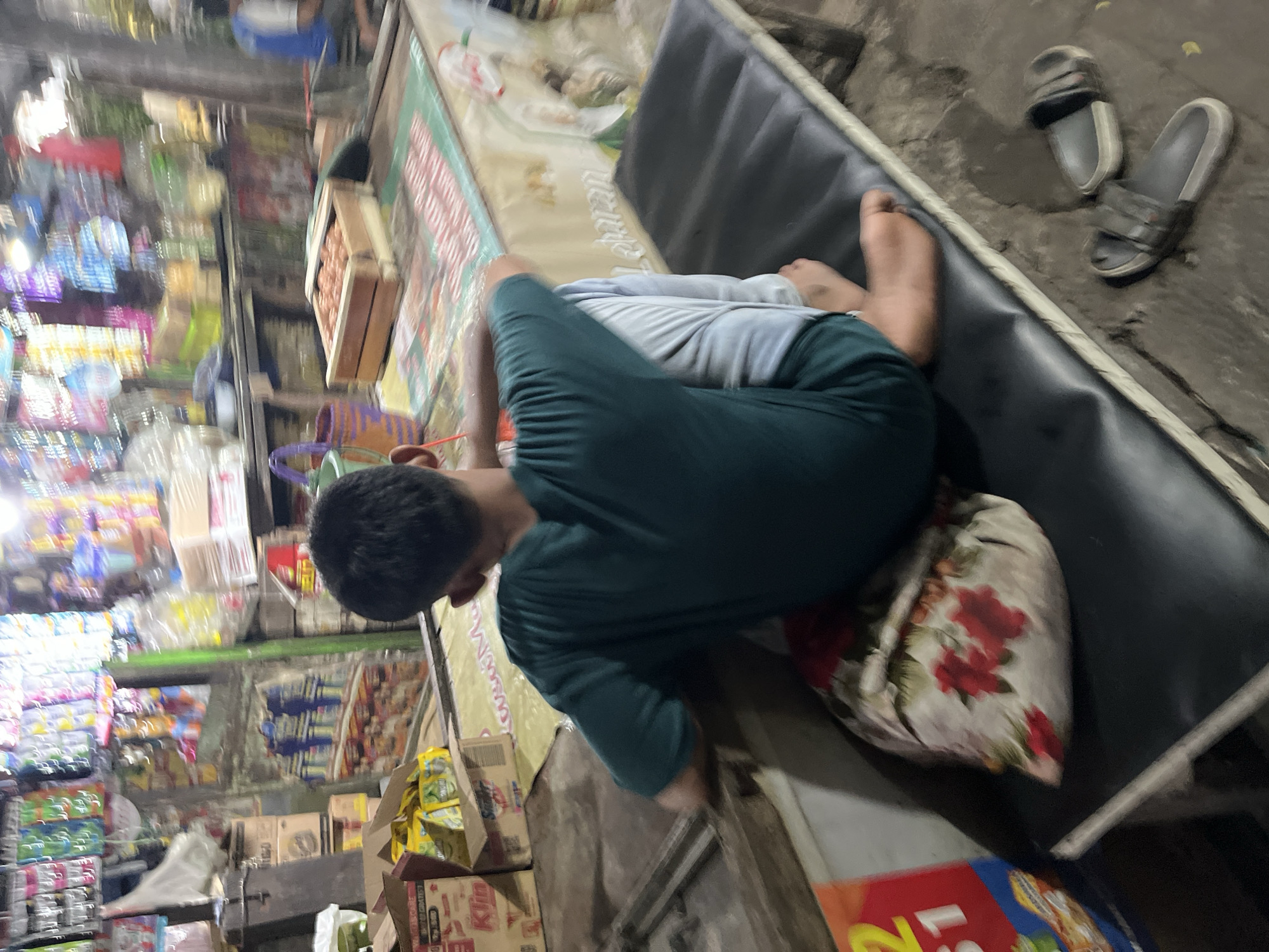 The image depicts a scene at a market stall in Larangan, Indonesia. A young person, viewed from the back, is seated on a dark-colored bench, resting their head on their knees. They are wearing a dark green t-shirt and light-colored pants. Underneath them is a pillow with a floral pattern featuring red roses. To the left of the bench, a portion of a red and yellow sign is visible, and further left, there are stacked cardboard boxes and yellow packaged goods.
The market stall itself is filled with various items. Behind the seated person, shelves are laden with colorful packaged goods and bottles. In the mid-ground, a wooden crate filled with small, round items, possibly eggs, is placed on a counter. The counter has a patterned mat or cloth on it with red and white stripes. The overall environment appears to be an open-air market, with dirt flooring visible beneath the bench. A pair of grey sandals rests on the ground near the right side of the bench. The lighting is somewhat dim, suggesting it might be late afternoon or evening.