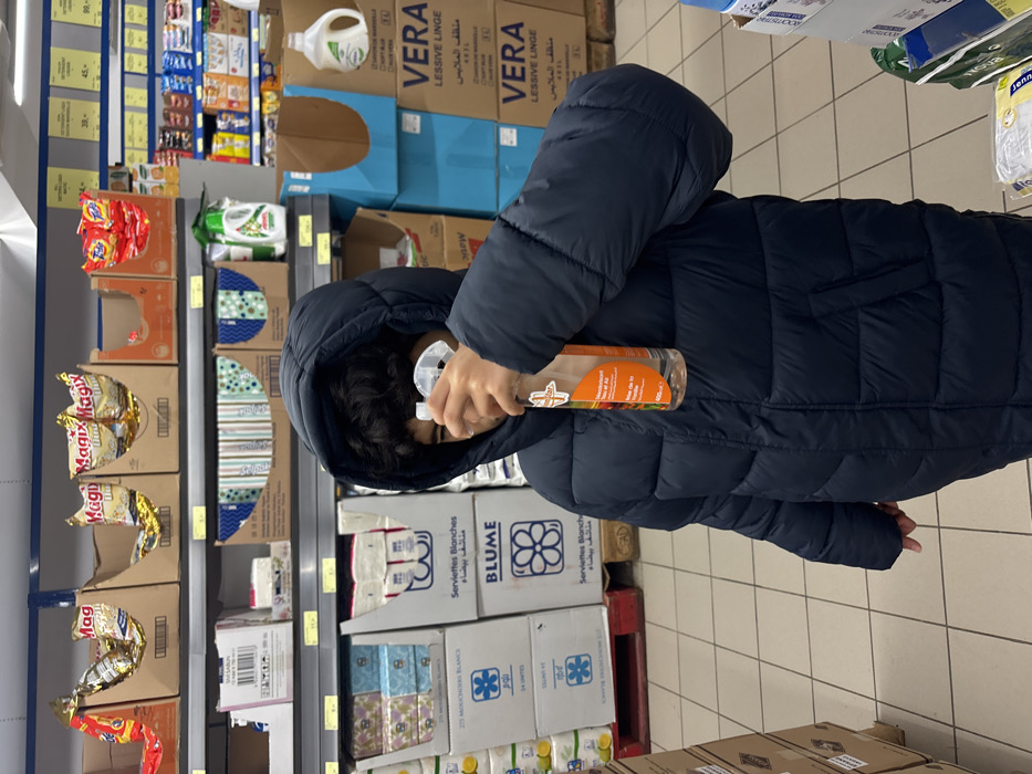 In a store aisle, a person in a puffy blue jacket sprays an orange bottle. Shelves hold a variety of products including "Magix" snack packs, patterned boxes, and white "Blume" tissues. Taller boxes of "VERA" laundry detergent in blue and brown are also visible. A shelf of assorted snacks and cleaning supplies is situated in the background. The floor is tiled.
