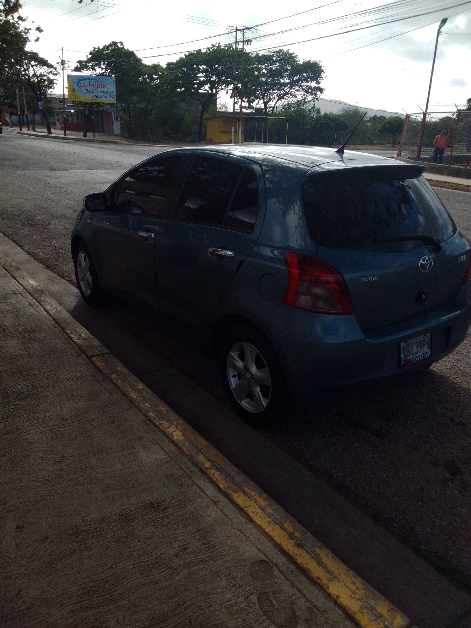 A blue Toyota Yaris is parked on the side of a road in Altagracia, Venezuela. The car is positioned diagonally, with its rear facing towards the right of the frame. The scene appears to be an urban or suburban environment, with trees and buildings visible in the background. A billboard advertising "carboline Recubrimientos Industriales" is present on the left side of the image. The sky is overcast, suggesting it may be a cloudy day. The time of day appears to be late afternoon or early evening, as indicated by the long shadows. The road surface is asphalt, and a concrete sidewalk with a yellow stripe runs alongside it. A person in an orange shirt is visible in the distance near a fence. The license plate of the car reads "ABZ39P4".