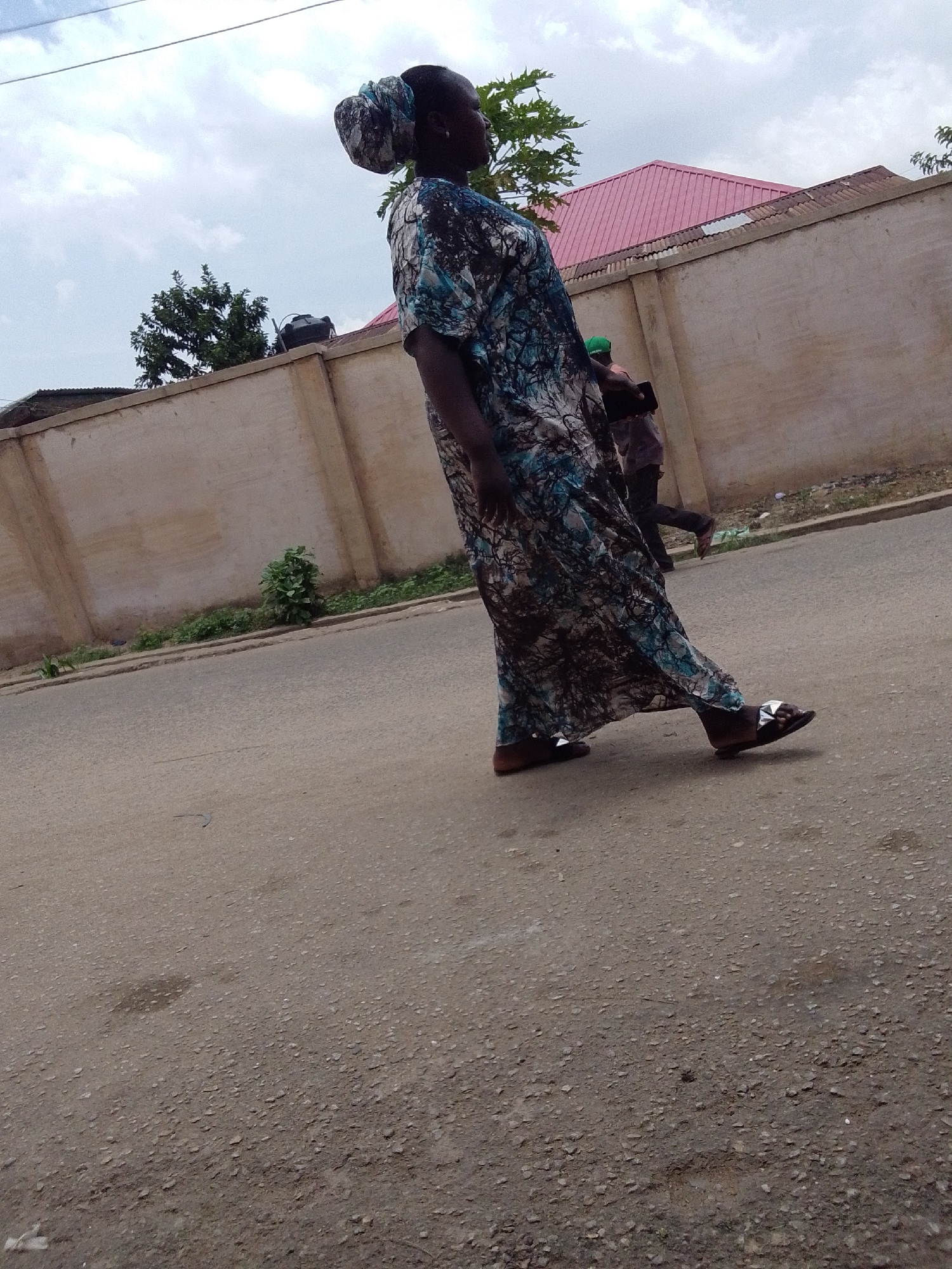 A woman wearing a long, patterned dress and sandals walks on an asphalt street in Jos, Nigeria. She has a patterned head covering. The street runs parallel to a concrete wall. Behind the wall, a building with a pink roof is visible, along with some trees. Another person is walking behind the woman, near the wall. The sky is overcast. The road surface is weathered.