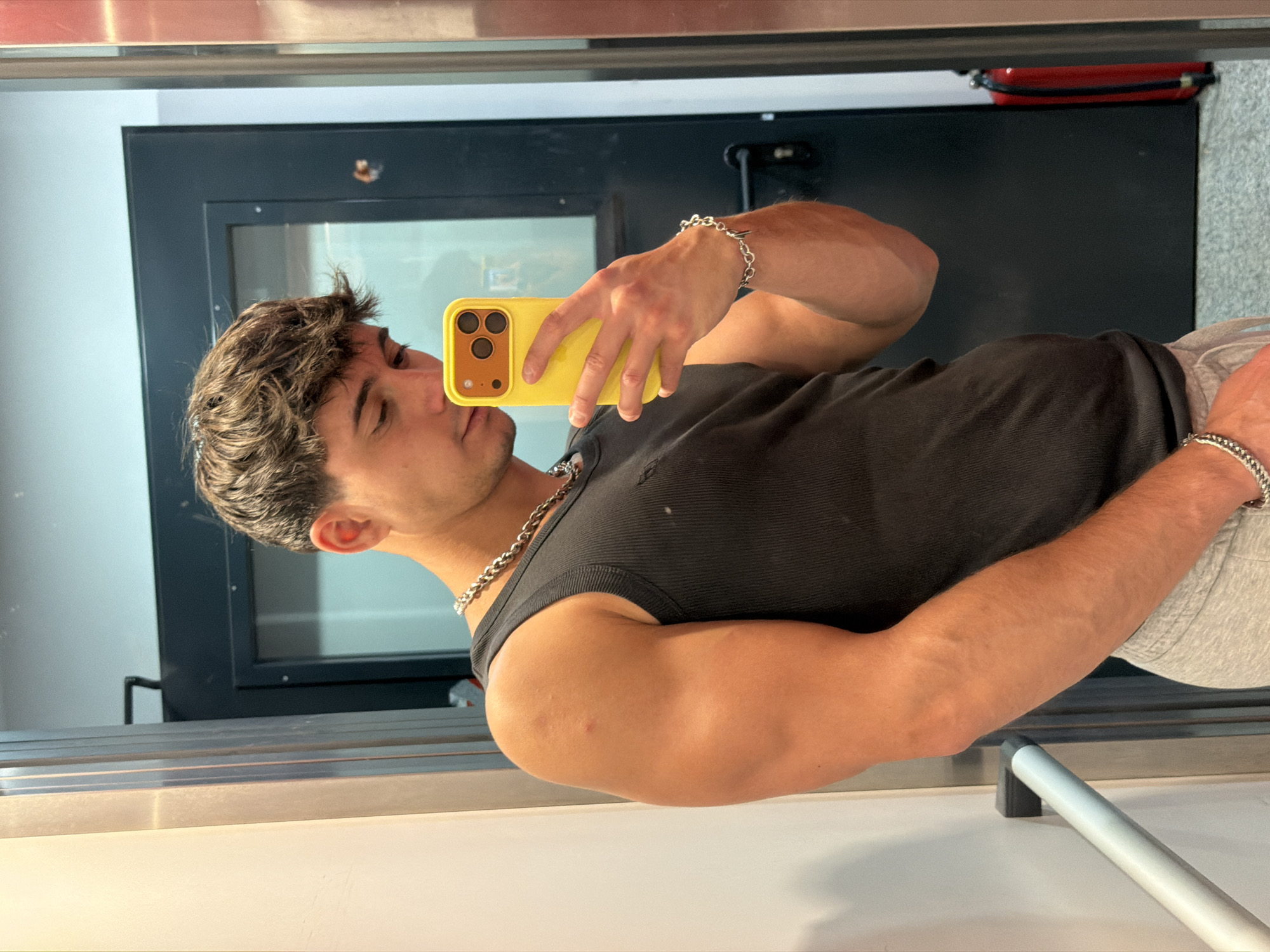 The image shows a young man taking a mirror selfie. He is wearing a black tank top, grey sweatpants, and silver chain necklaces and bracelets. He is holding a yellow smartphone with a case in his right hand. The man appears to be in an indoor environment, possibly a gym or locker room, with a metal door and a fire extinguisher visible in the background. The flooring appears to be carpeted. There is no visual information in the image that can be used to determine the location within the city of Sofia, Bulgaria.