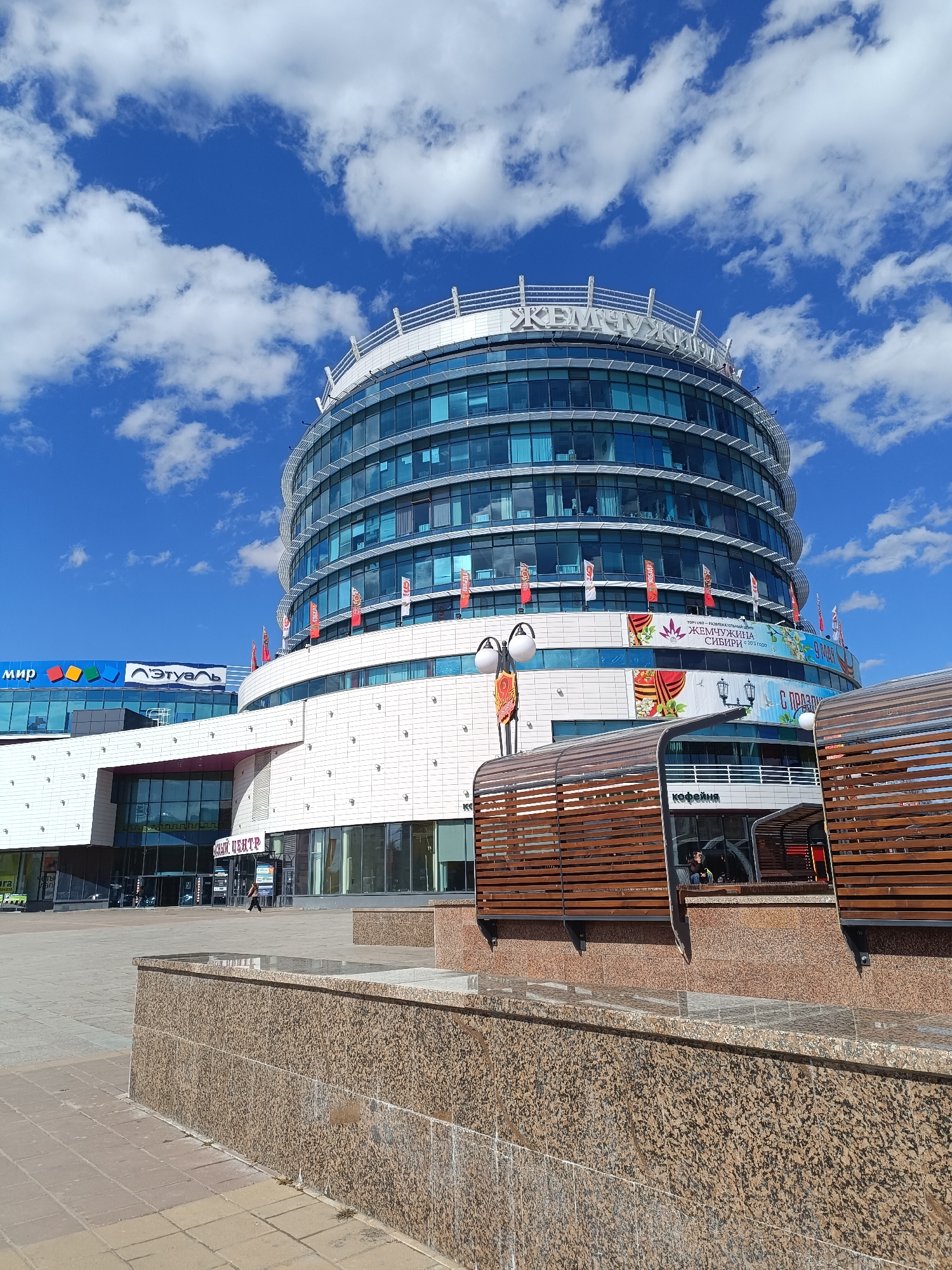 This image captures a modern, multi-story building complex in Tobolsk, Russia, under a bright blue sky with scattered white clouds, indicating a clear daytime. The central and most prominent structure is a large, circular building with a reflective blue-tinted glass facade on its upper floors and white tiled walls on the lower levels. The building is topped with a white railing and features large, white Cyrillic letters spelling "ЖЕМЧУЖИНА" (ZHEMCHUZHINA), meaning "Pearl," on its roofline.
Numerous small flags in red, white, and other colors are affixed to the facade of the round building. A large banner on one of its lower levels reads "ТОРГОВО-РАЗВЛЕКАТЕЛЬНЫЙ ЦЕНТР ЖЕМЧУЖИНА СИБИРИ С 2003 ГОДА" (TRADE AND ENTERTAINMENT CENTER PEARL OF SIBERIA SINCE 2003), along with "9 МАЯ" (9 MAY) and "С ПРАЗДНИКОМ" (HAPPY HOLIDAY), suggesting a celebration related to Victory Day.
To the left, an adjacent building, also modern with glass and white panels, displays signs for "мир" (Mir), identifiable by a colorful square logo, and "Этуаль" (L'Etoile), a well-known cosmetics and perfumery chain.
In the foreground, there's a paved pedestrian area with granite-clad low walls or planters. Behind these walls are contemporary outdoor seating structures made of dark wood slats. One of these structures partially obscures a sign for a "кофейня" (coffee shop). A lone person can be seen walking across the paved area further back, near an entrance marked with "ЦЕНТР" (CENTER). The overall scene depicts a bustling, commercial urban environment.