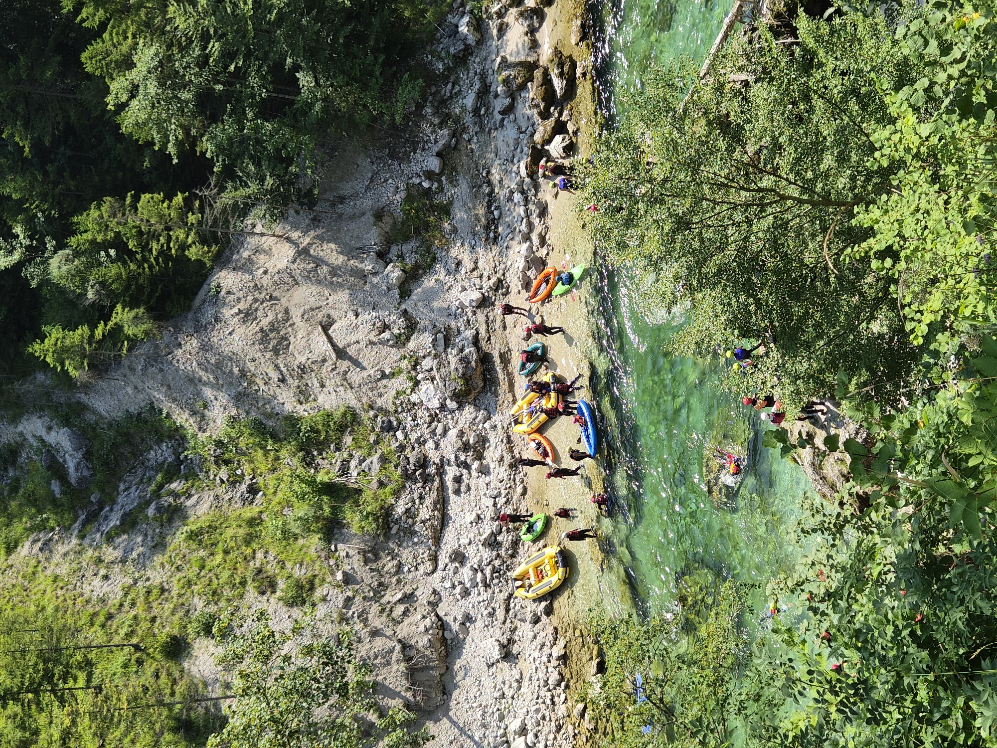 In the immediate foreground, lush green foliage obscures the bottom portion of the image. The midground depicts a group of people and several inflatable rafts and kayaks on the rocky bank of a clear, emerald-green river. The people are wearing life vests and helmets, and some are actively engaged in launching or preparing the watercraft. The riverbank is composed of pebbles and larger stones, with a steep, vegetated slope rising behind it. The background features a dense forest of green trees on a hillside under a bright, sunny sky. This scene is located in the city of Landl, Austria, specifically on the Enns River, known for its white-water rafting and kayaking activities.