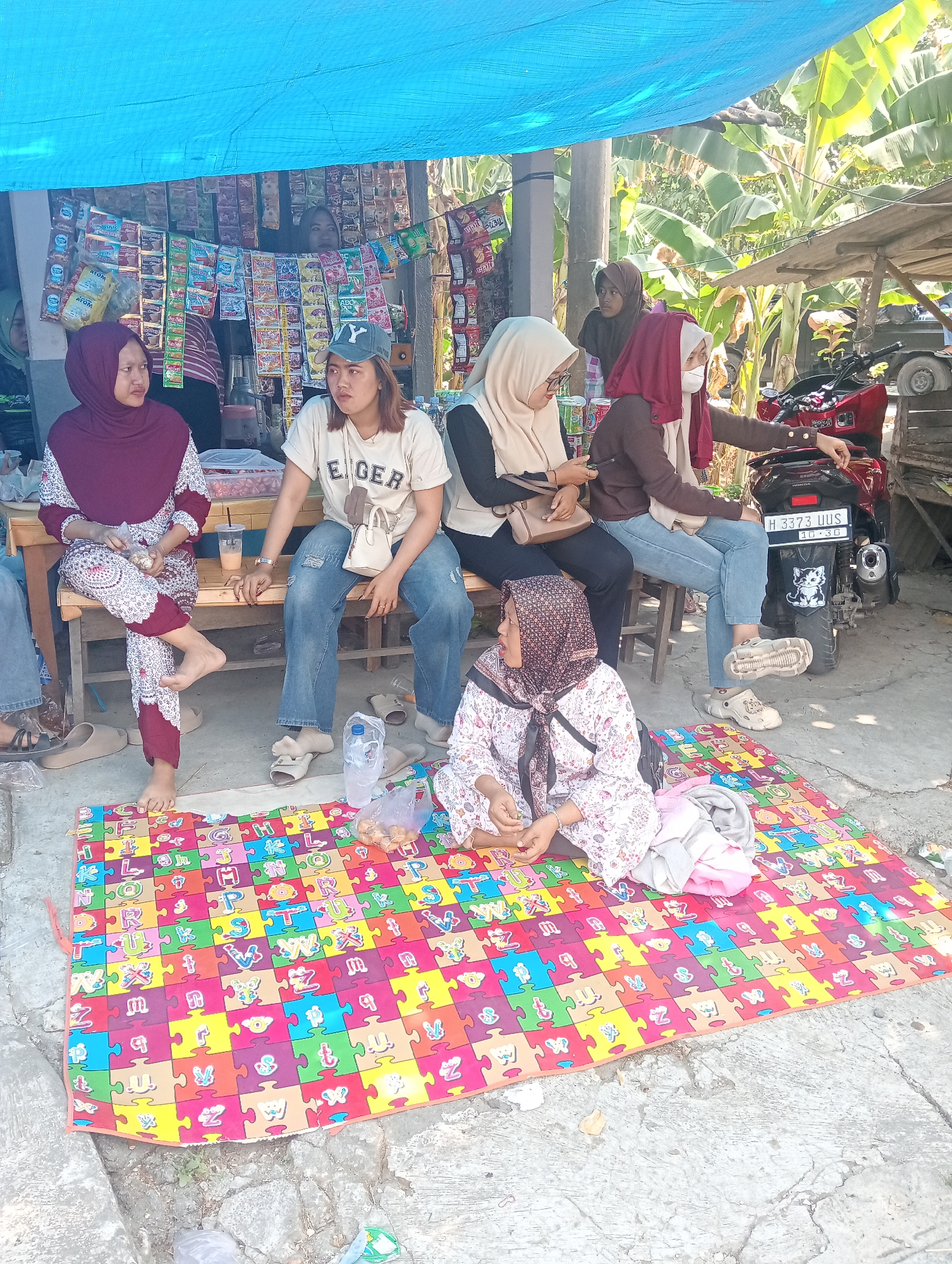 The image depicts an outdoor scene in Doreng, Indonesia, under a blue tarp. In the foreground, a colorful alphabet puzzle mat is spread on the ground. Several people are present. A woman wearing a maroon hijab and patterned pants sits on a wooden bench to the left, holding some snacks. Next to her, a woman in a white t-shirt and blue jeans, wearing a blue baseball cap, also sits on the bench. Another woman in a beige hijab is seated on the bench, looking at her phone. To the right, a woman in a maroon hijab and a brown jacket is seated on a chair, pointing towards a red motorcycle parked behind her. A woman in a patterned hijab and white floral dress is seated on the puzzle mat in the foreground, looking to her left. A small shop with various packaged goods is visible in the background. The environment suggests a casual gathering or a small roadside stall.