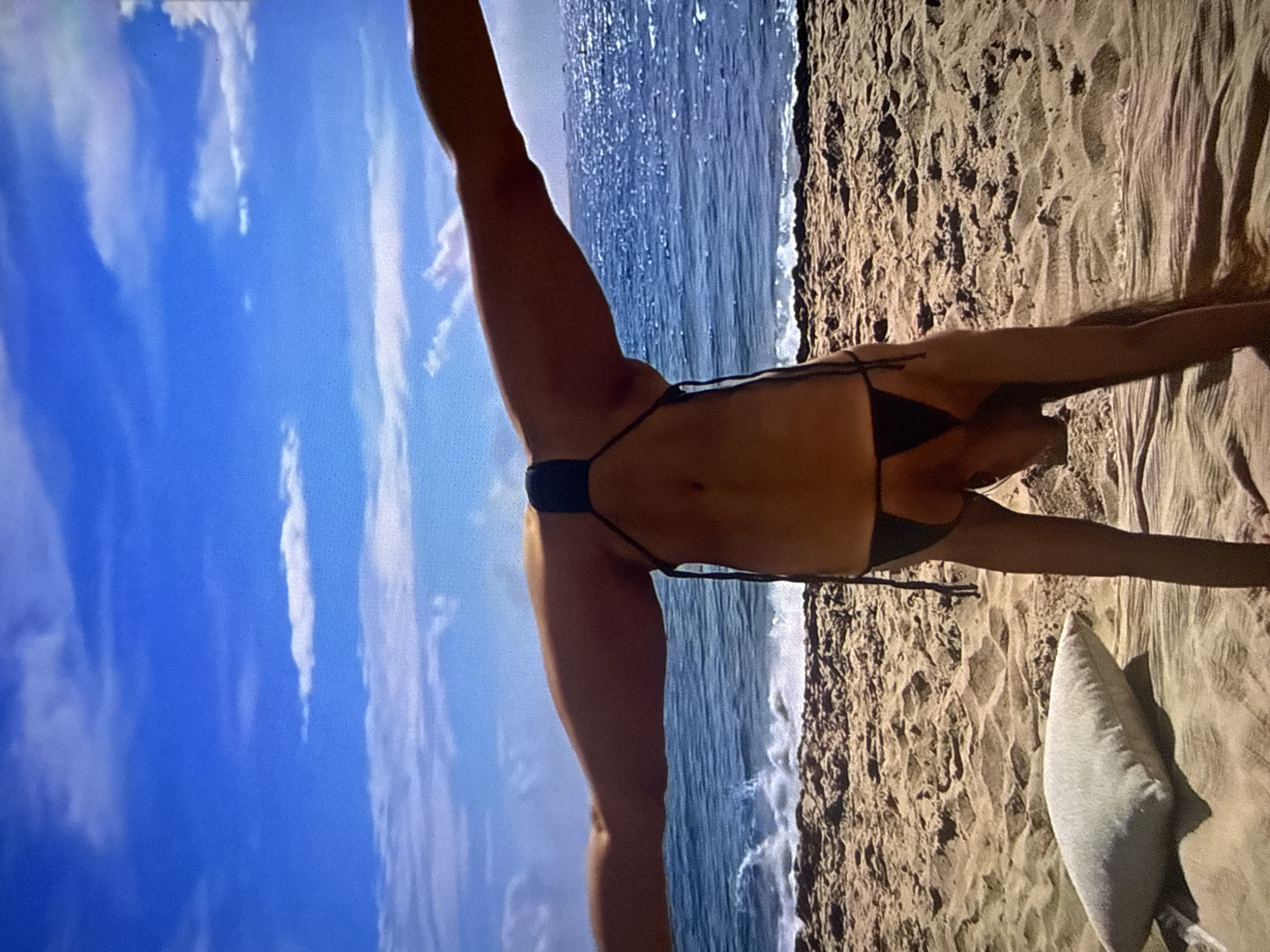 The image displays an outdoor scene on a sunny beach, stated to be located in Varna, Bulgaria. A single individual, appearing to be female, is positioned centrally in an inverted acrobatic or yoga pose. The person is performing a handstand with their legs widely splayed upwards and outwards in a V-shape. They are wearing a black string bikini. The individual's head is oriented downwards towards the sandy ground, and their face is not visible.
The background features a bright blue sky with scattered white, wispy clouds. The sky meets a deep blue ocean horizon, where several large vessels or ships are faintly visible in the distance, particularly in the upper right quadrant of the water. White-capped waves are breaking near the shoreline. The foreground consists of light golden-brown sand with visible ripples and disturbances. A light-colored, rectangular pillow rests on a darker, possibly fabric, surface on the sand to the left of the person's lower body.