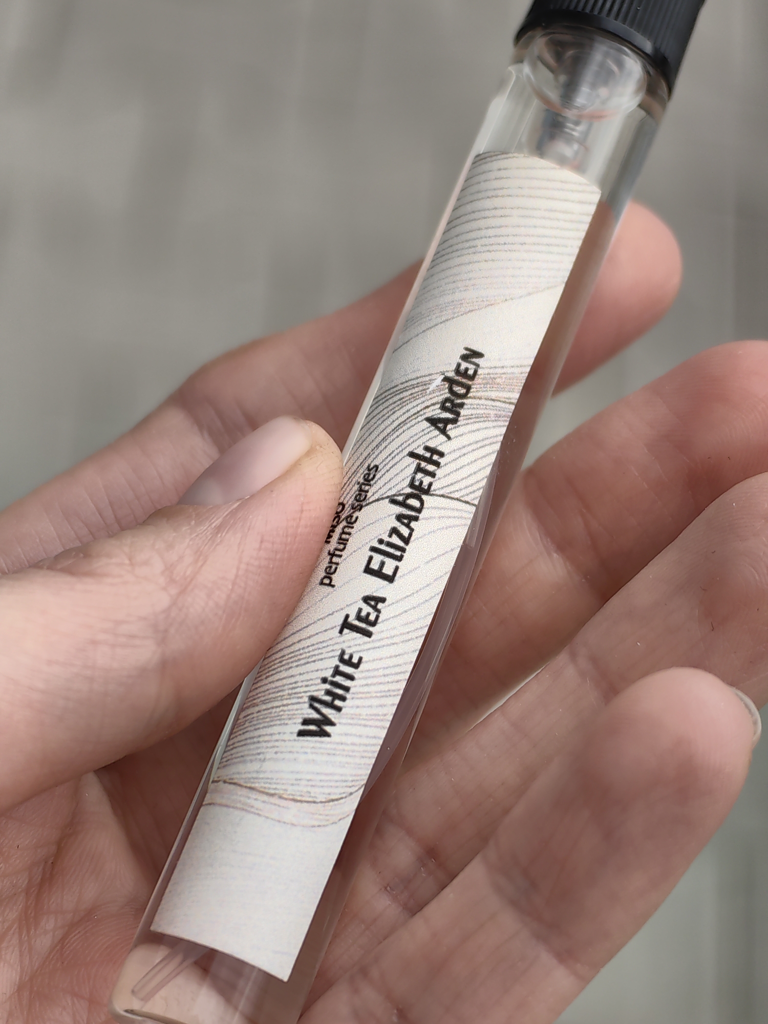 A close-up, eye-level shot shows a person's left hand holding a small, clear glass perfume sample vial. The hand, with light skin tone, occupies a significant portion of the bottom and left side of the frame, with visible fingers and a thumb. The background is a soft, out-of-focus light gray, suggesting an indoor setting or diffuse natural light.
The central object is the slender glass vial, approximately 2-3 inches long, with a black screw-on cap at the top. The vial contains a clear liquid and an internal spray mechanism. A white label with fine, light gray, wavy horizontal lines is wrapped around the middle of the vial. Black text is printed on this label.
Visible text on the label reads: "perfume series" (partially visible, positioned vertically) and "White Tea Elizabeth Arden" (prominently displayed horizontally and slightly angled). The label appears to have a slightly textured or patterned background. The activity is simply presenting or holding this perfume sample. There are no specific location or time-of-day cues beyond the diffuse lighting. No emotions are discernible. The image captures the details of the vial and the hand holding it.