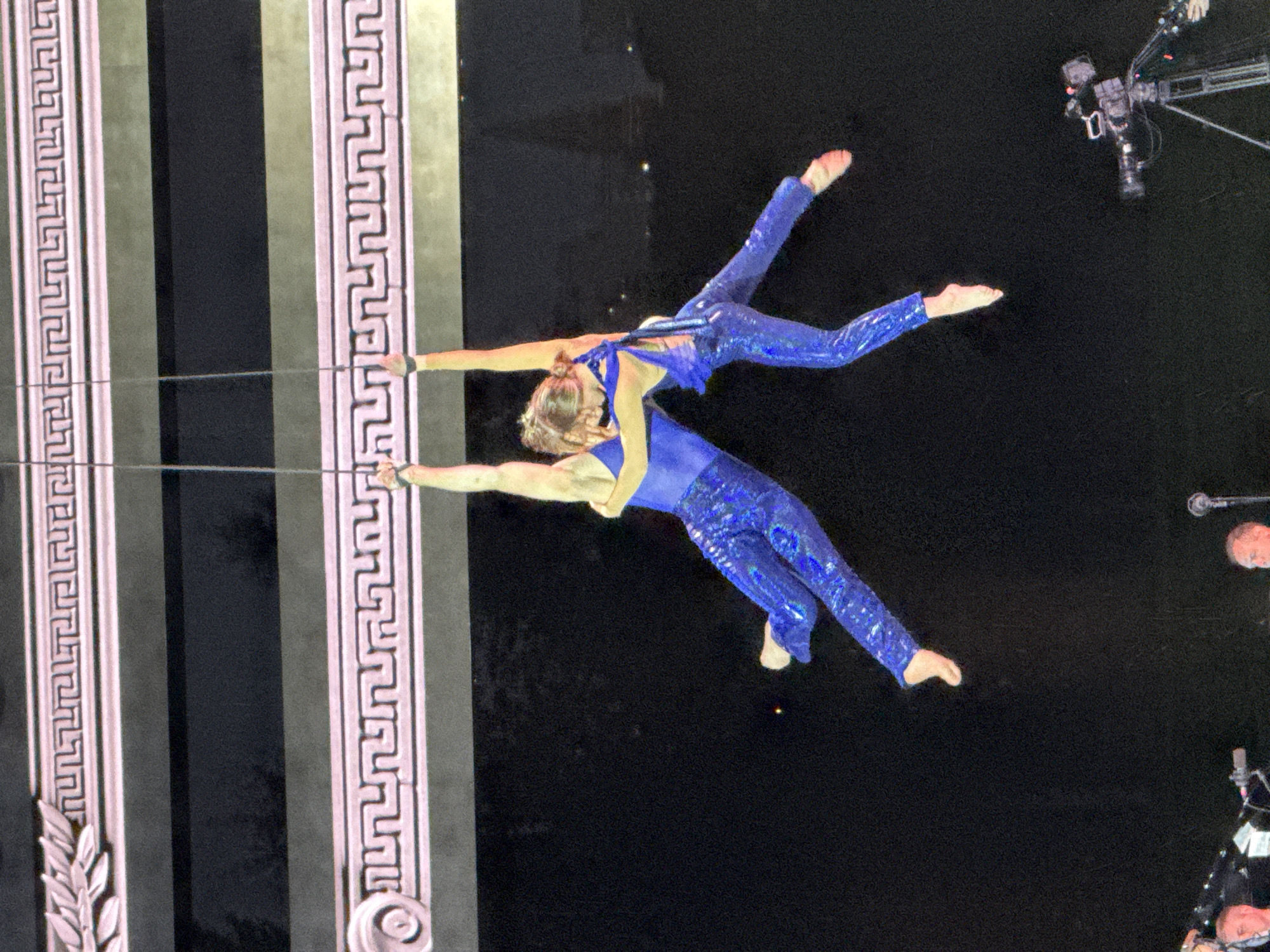 A duo of aerialists, clad in sparkling blue costumes, perform on ropes suspended from an ornate ceiling structure. The aerialists are in mid-air, engaged in a dynamic pose, with one holding onto the other's hands and legs. The background is a dark, textured surface, likely a stage curtain or wall. Below, in the foreground, are elements of a stage setup, including a camera on a tripod, and parts of musical instruments or sound equipment. The lighting appears to be stage lighting, creating a dramatic effect.