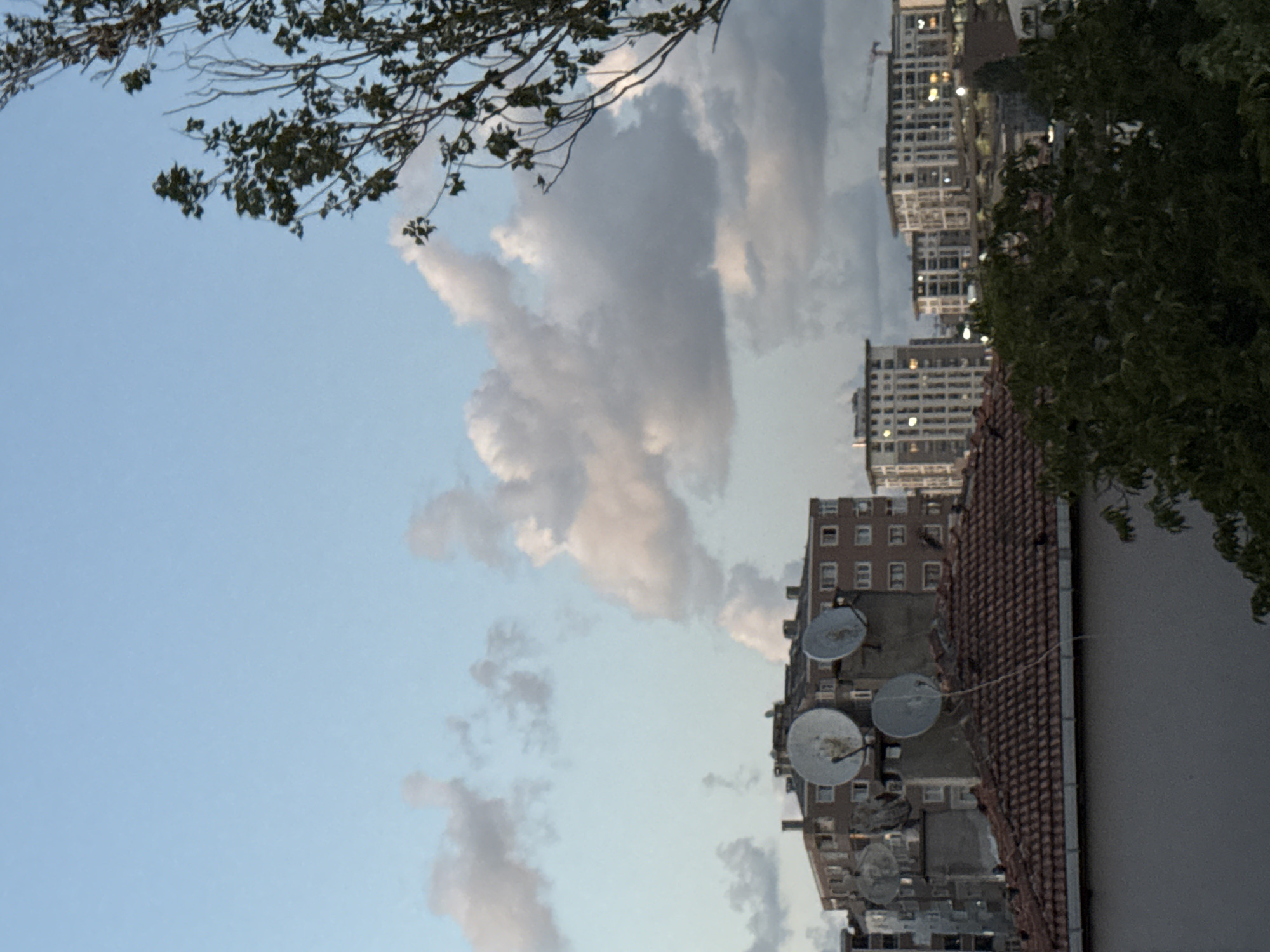 The image depicts a cityscape in Gungoren, Turkey, under a sky with scattered clouds. In the foreground, a brown-tiled roof of a building is visible. Above this, a multi-story building with many windows and satellite dishes mounted on its exterior is prominent. Further back, several modern apartment buildings with lit windows rise against the sky. A crane can be seen in the distance to the right, suggesting ongoing construction. Trees with green foliage are present in the lower right corner of the image. The overall scene is a snapshot of urban life with residential buildings and clear sky.