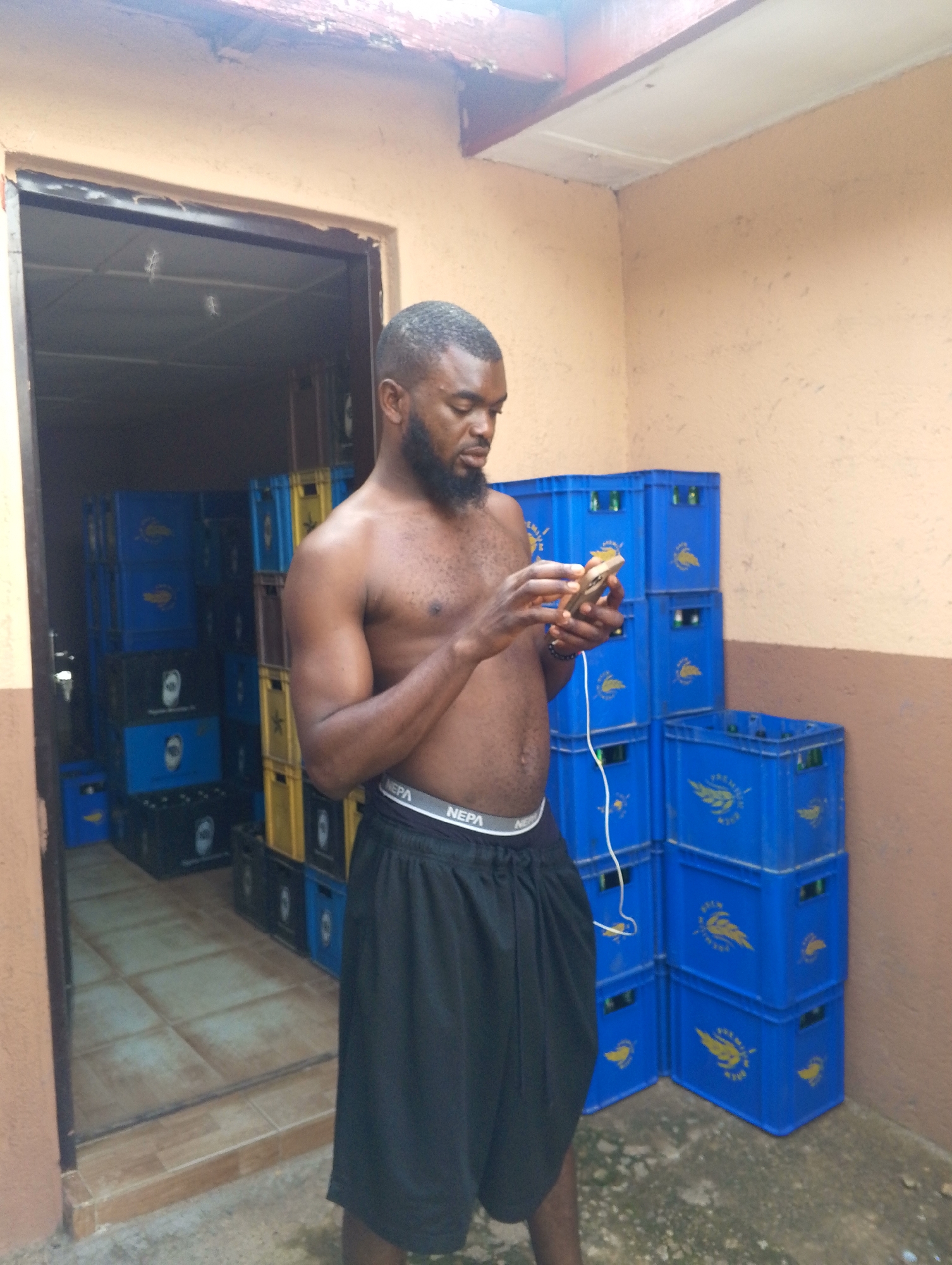A shirtless man with a beard stands in front of a doorway, looking at his phone. He is wearing black shorts with a "NEPA" logo visible on the waistband. Behind him, inside the doorway and to his right, are stacks of blue plastic crates, many filled with bottles. The crates have a yellow winged logo and text that appears to say "PREMIER BEER". Some yellow and black crates are also visible. The scene appears to be outdoors, with a stucco wall to the right and a tiled floor inside the doorway. The time of day seems to be daytime, possibly afternoon, given the lighting. The man's expression is focused on his phone. This appears to be a storage area or a small shop in Jos, Nigeria.