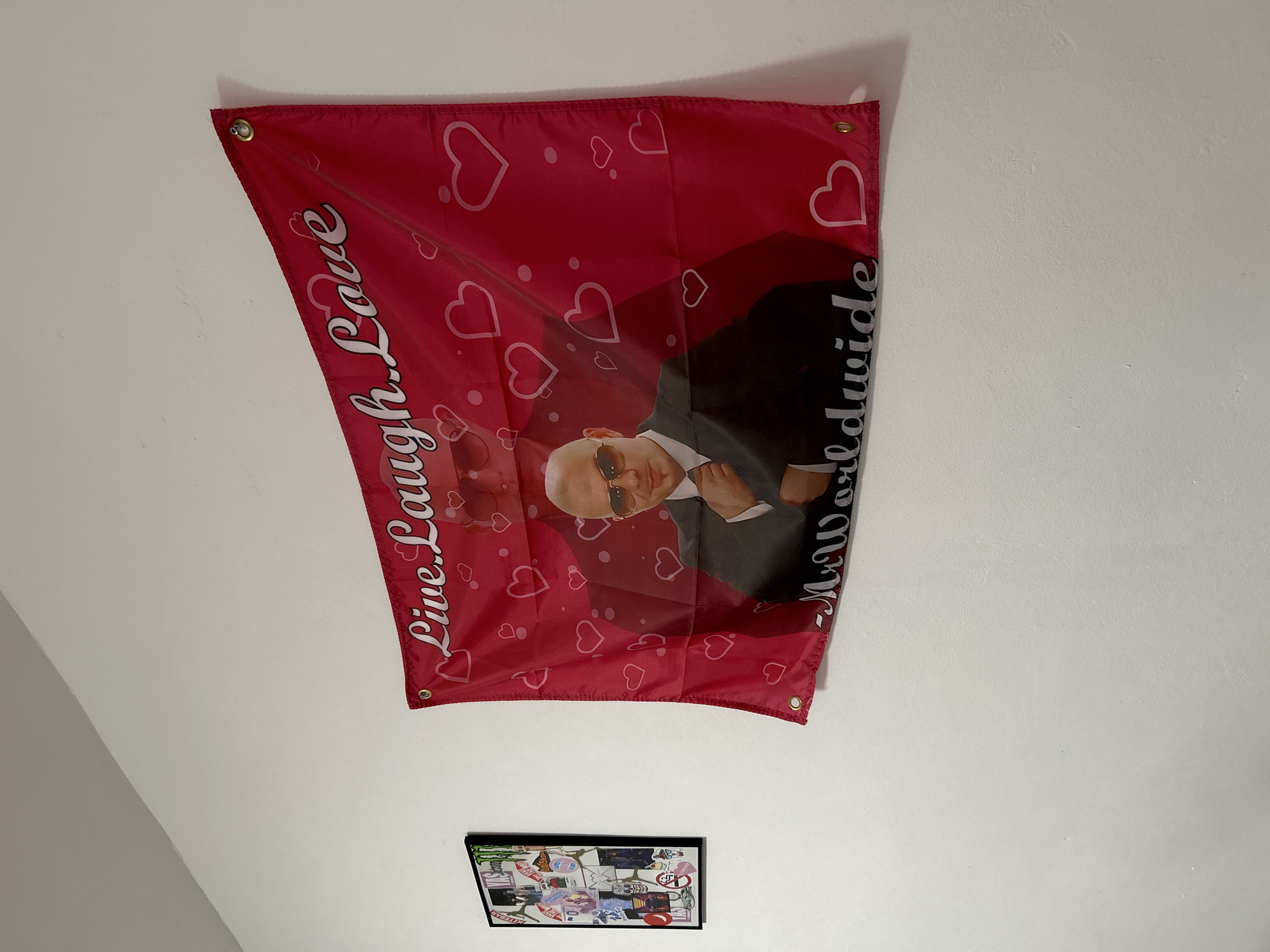 The image depicts a wall with two items hanging on it. The upper item is a red flag with a photo of Pitbull, the rapper, wearing sunglasses and a suit, against a background of white hearts. The flag also says "Live, Laugh, Love - Mr. Worldwide" in white lettering. Below the flag is a framed picture with numerous stickers on it. The wall is white. The location is not discernable from the image.