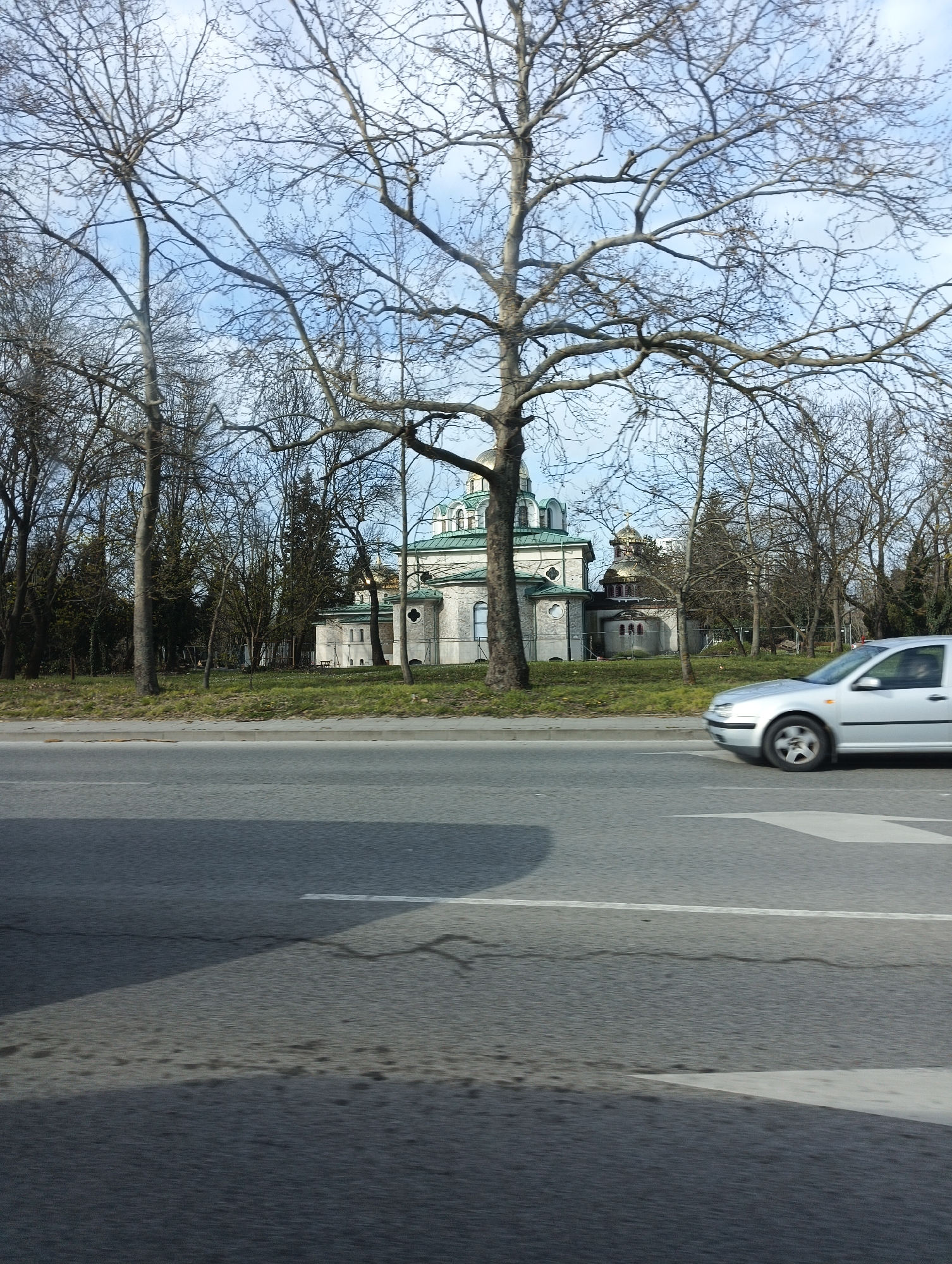 The image shows a view across a road towards a light-colored building with green domes, likely a church, partially obscured by bare trees. A silver car is moving on the road, creating a blurred effect. The road is asphalt, with a white line marking the lanes. The sky is pale blue with some clouds.
Given the architecture and appearance, the church is likely located in Burgas, Bulgaria. Specifically, it could be the church "St. Cyril and St. Methodius".