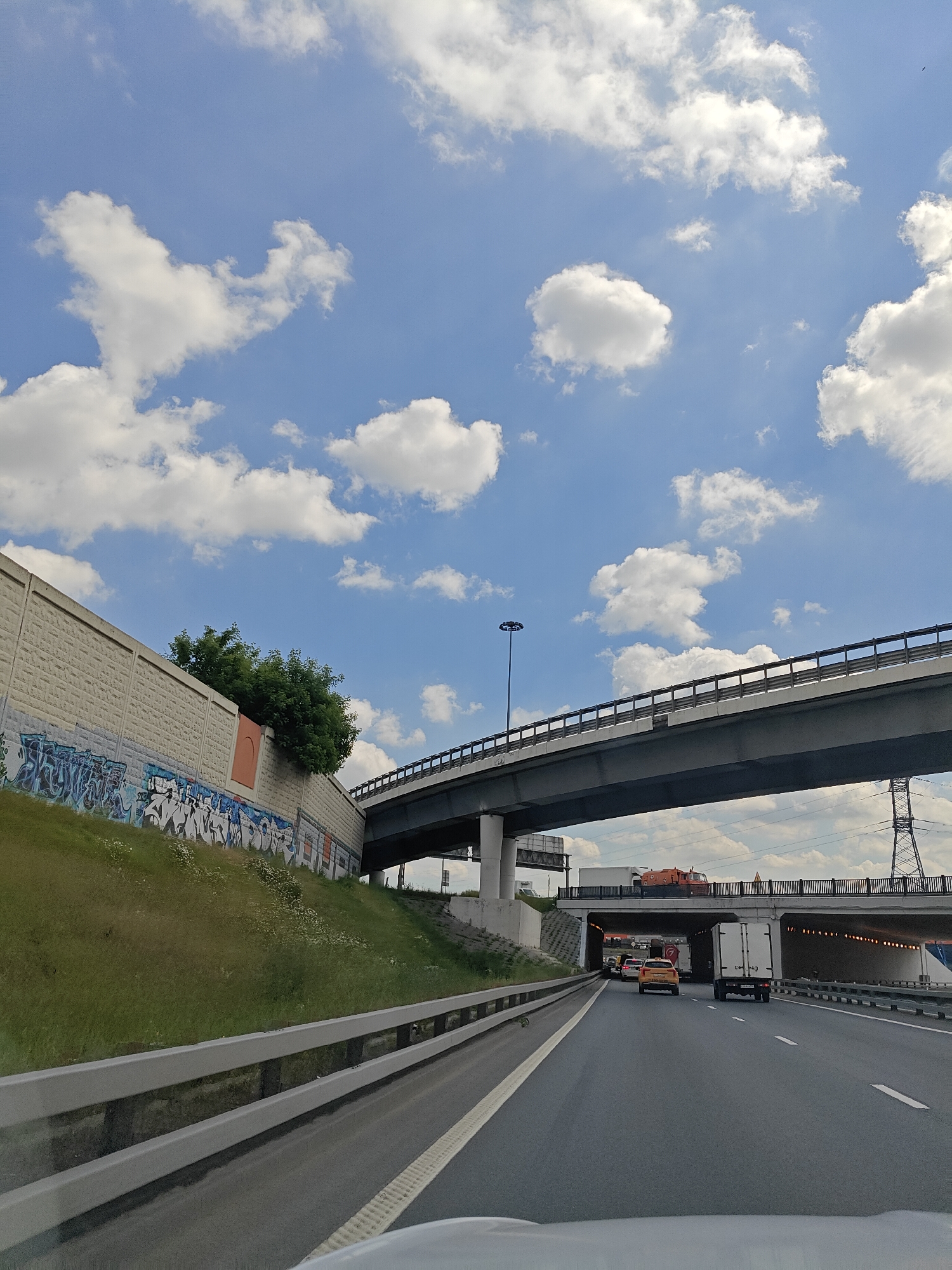 The image shows a view from a car driving on a multi-level highway in Mytishchi, Russia. The sky is blue with scattered white clouds. A concrete retaining wall with graffiti is on the left, covered in grass. Above the retaining wall, a large elevated highway structure is visible. Below this structure, another highway is in view with several vehicles, including cars and trucks. There are power lines and pylons in the background.