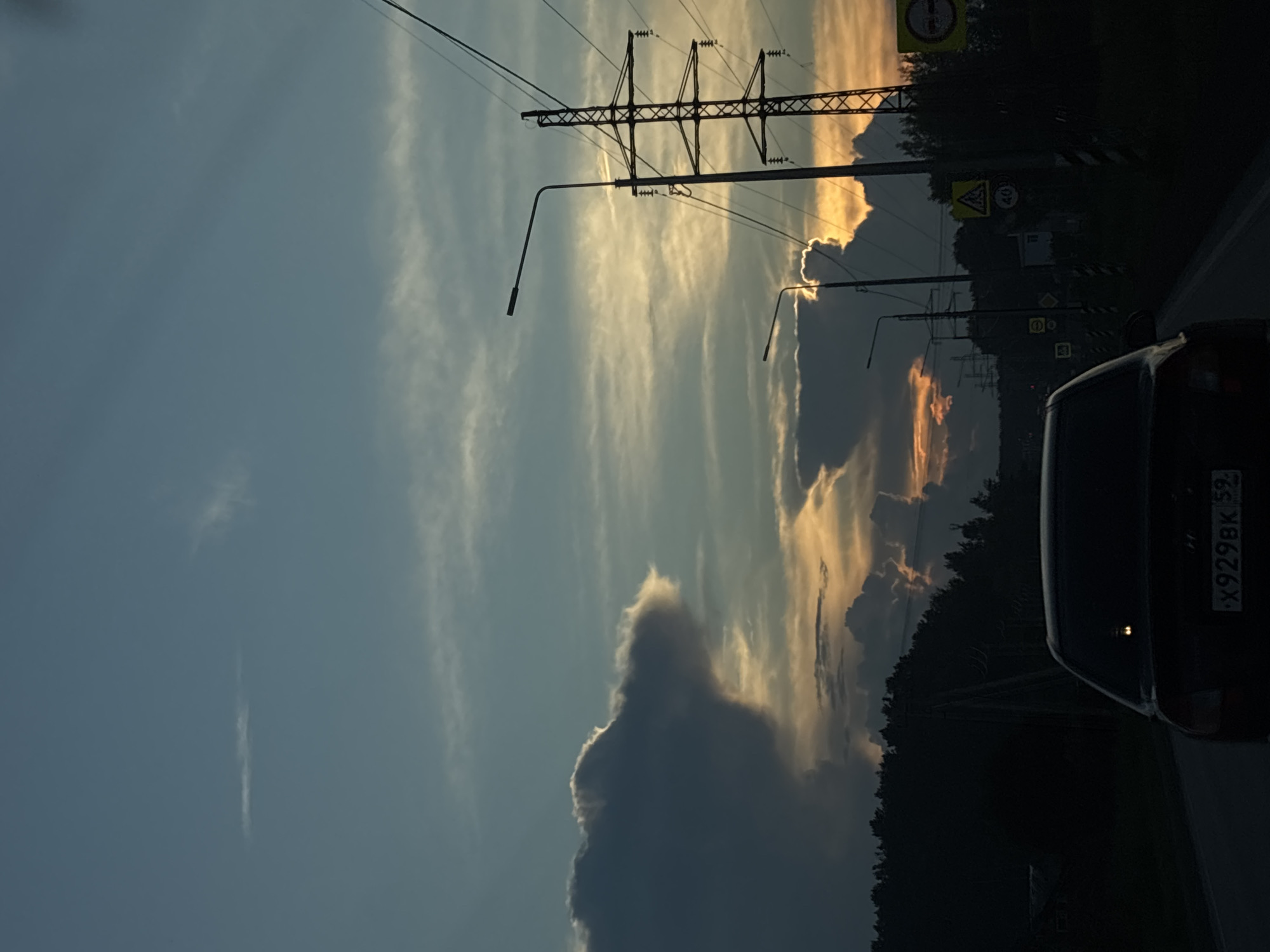 The image depicts a road view from inside a vehicle, likely at dusk, with a dark sedan in the foreground. The sky is a mix of blue and pale orange hues, with scattered clouds. Large, dark clouds dominate the left side of the frame, while lighter clouds are visible near the horizon, illuminated by the setting sun. Electrical pylons and streetlights line the right side of the road. Trees and foliage form a dark silhouette along the left horizon. Several road signs are visible on the right side of the road. The license plate of the vehicle in the foreground is partially visible and reads "X929BK 59". The scene appears to be on a road in or near Chaikovsky, Russia, given the presence of the regional code "59" on the license plate, which corresponds to the Perm Krai region where Chaikovsky is located.