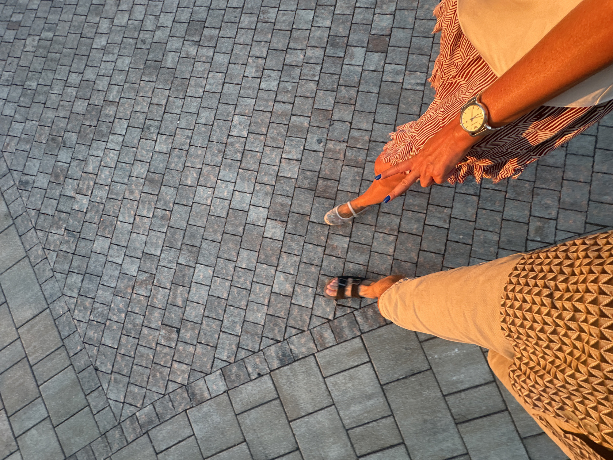 The image shows a high-angle perspective of two people walking on a stone-paved area in Kumbor, Montenegro. The paving consists of rectangular grey stones, arranged in a pattern with a slight curvature. Sunlight casts long shadows on the ground, indicating it is either early morning or late afternoon.
On the left, one person's legs are visible wearing light-colored, loose-fitting trousers with a geometric patterned trim near the ankle. They are wearing dark sandals.
On the right, another person's legs are visible, one in focus and the other slightly blurred. This person is wearing a white sandal on their left foot and a red and white striped dress or skirt. Their right hand, adorned with a silver wristwatch and bright blue nail polish, is visible in the frame, with their index and middle fingers extended in a peace sign. The skin tone of this person appears to be tanned. The interaction suggests they are walking together. The environment is an outdoor urban setting.