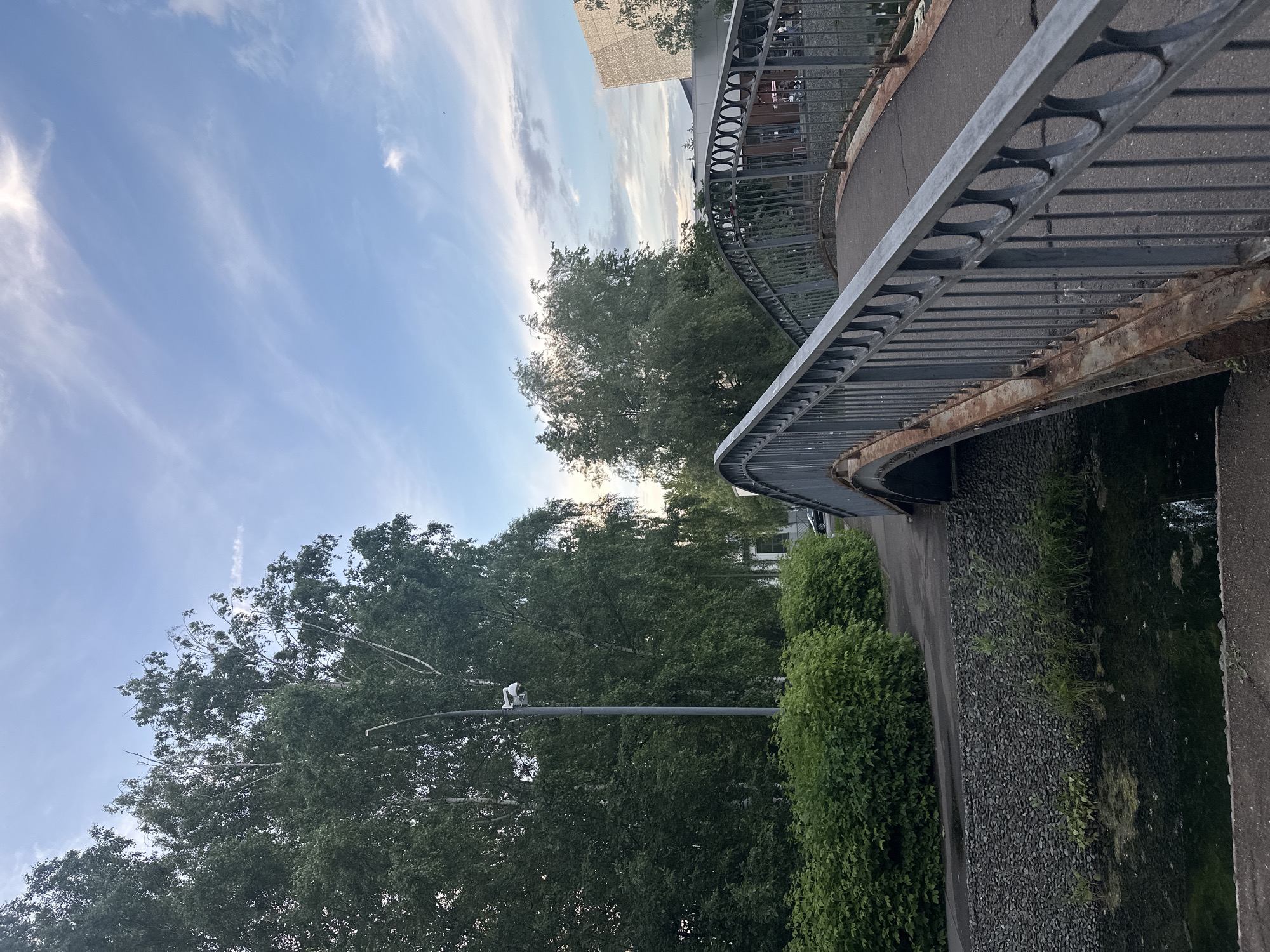 The image shows an arched pedestrian bridge with a metal railing, extending over a narrow, dark body of water. The railing is grey and shows signs of rust. The bridge's surface is paved. On the left side of the bridge, there is a stone embankment lined with lush green bushes and trees. A lamppost with a security camera is visible among the trees. A paved walkway runs alongside the embankment, and a black car is parked on the road in the background. On the right side of the bridge, a modern building with a geometric facade can be seen. The sky above is blue with scattered clouds. The scene appears to be an urban park or a green area within a city. Based on the architectural style of the modern building and the overall environment, this location is likely the park area surrounding the Kazan Family Center, also known as "The Bell", in Kazan, Russia.