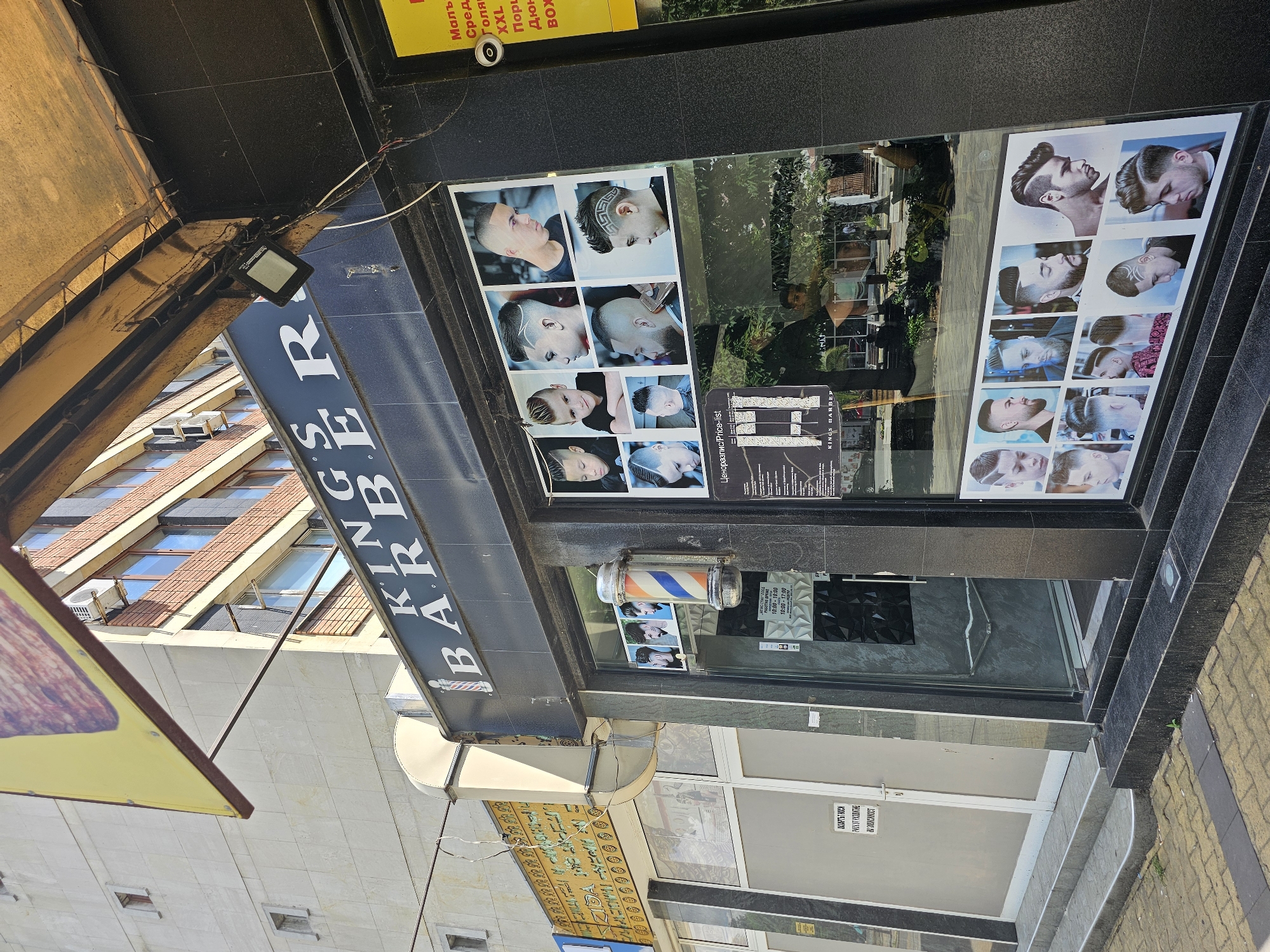 The image shows the exterior of a barbershop called "KING'S BARBER." The shopfront is decorated with photos of various men's hairstyles. A traditional barber's pole is mounted on the wall. Signs with text in both Bulgarian and English can be seen. A building can be seen in the background, suggesting an urban environment. The location is Karnobat, Bulgaria.