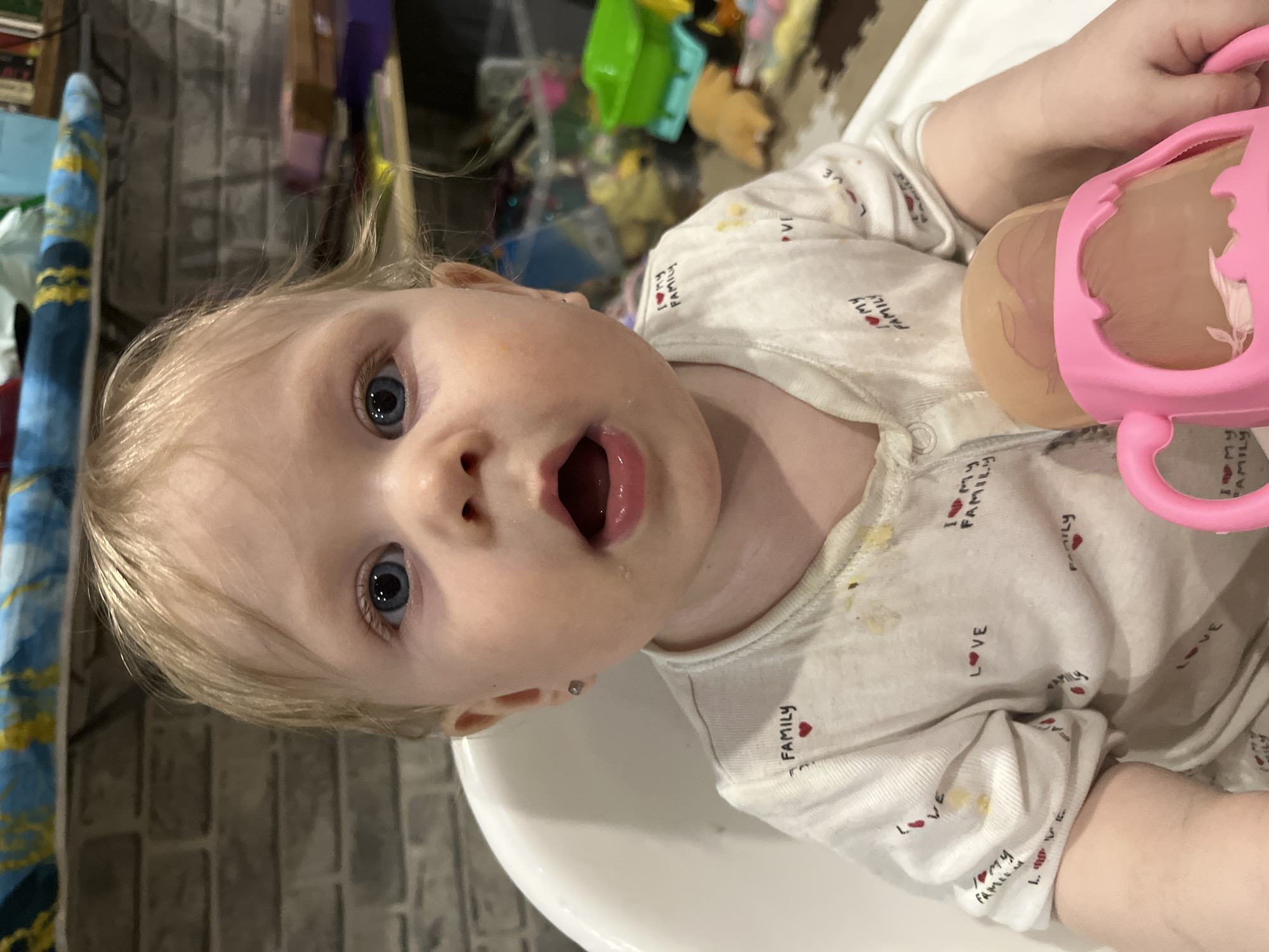 A fair-skinned baby with blonde hair and blue eyes sits in a high chair, looking up with an open mouth. The baby is wearing a light-colored onesie patterned with red hearts and the text "I ❤️ MY FAMILY" and "LOVE." The onesie has some yellow stains on it. The baby is holding a pink sippy cup in their right hand. The background is a cluttered indoor space with toys and other items. A striped blue and gold object is visible to the left of the baby's head. The location appears to be indoors, possibly a kitchen or dining area. No information suggests this image was taken in Vesyoloye, Russia.