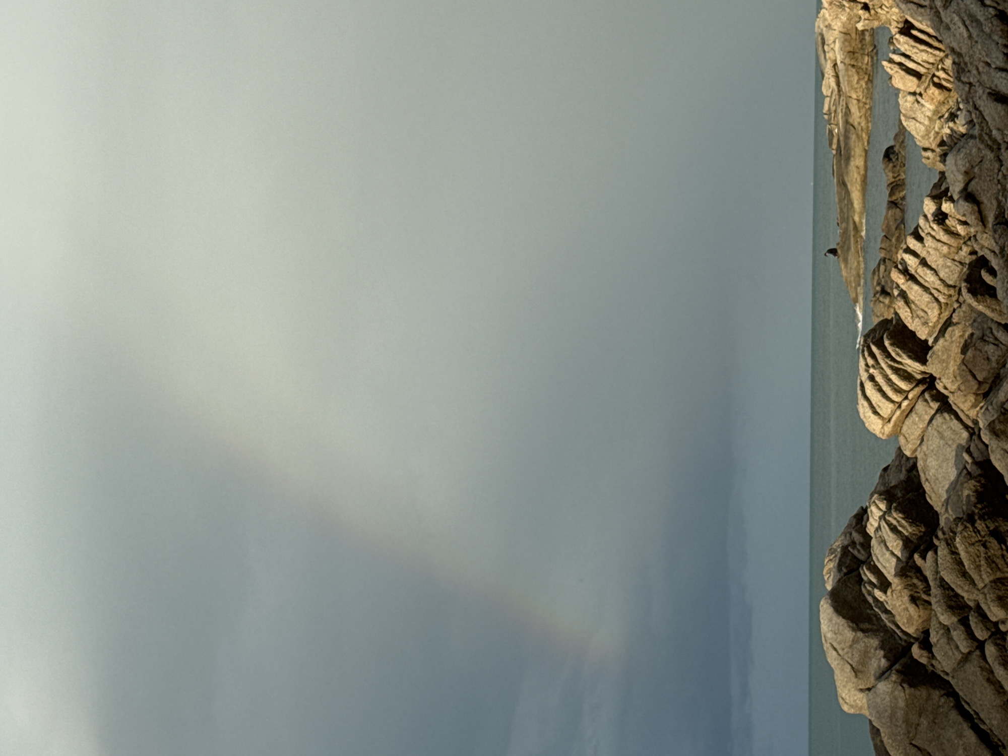 A faint rainbow arc is visible in a cloudy sky over the ocean, with a person sitting on a rock in the distance. The foreground is dominated by a rocky coastline with large, textured boulders. The water is calm, and the overall atmosphere is subdued due to the overcast conditions.