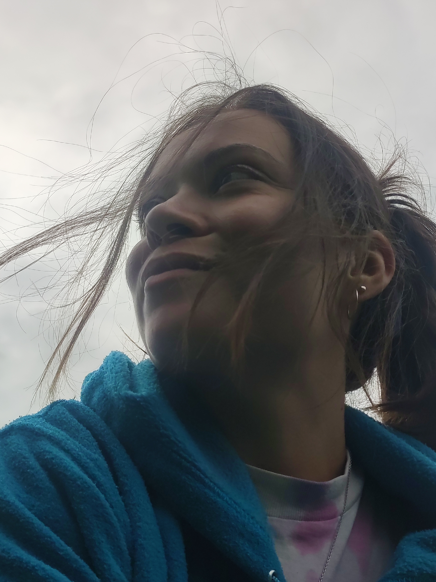 This is a close-up, low-angle outdoor shot of a young person, likely a female, from the shoulders up. The subject has brown hair that is windblown, with strands reaching across the frame. They are wearing a bright blue, textured hooded garment over a white t-shirt with a tie-dye pattern of pink and purple. A delicate silver chain is visible around their neck. The subject is looking upwards and to the right, with their left eye slightly visible. The background is a cloudy, overcast sky. The image does not contain any specific identifiable landmarks or features that would place it within the city of Фоки, Russia.