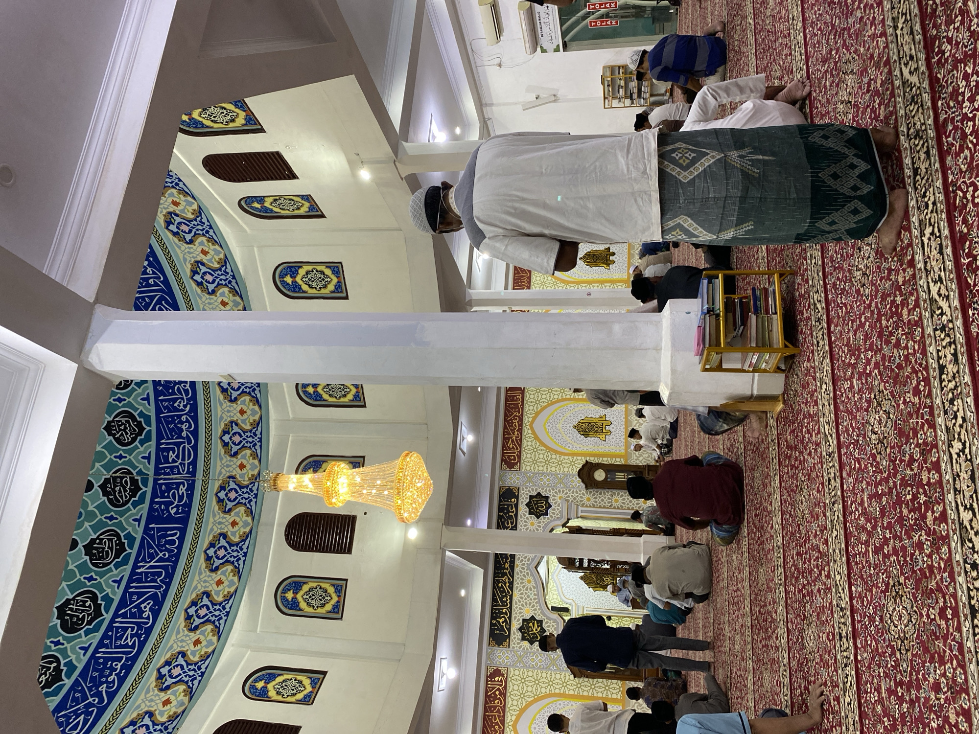 The image depicts the interior of a mosque, likely located in Lhokseumawe, Indonesia, given the context. Several Muslim men are present, some standing and facing the Qibla, others seated on a red carpet with intricate patterns. The mosque features a large, ornate chandelier hanging from the domed ceiling, which is decorated with Islamic calligraphy and colorful geometric patterns. Arched windows with stained-glass inserts are visible along the upper walls. A large white pillar is centrally located in the foreground, partially obscuring the view of some worshippers. The walls are adorned with decorative panels featuring Arabic script and geometric designs. A small yellow shelf unit filled with books is situated near the pillar. In the background, through an archway, a mihrab and minbar are visible, indicating the prayer direction and the elevated pulpit. The overall atmosphere suggests a religious gathering, possibly during prayer time.