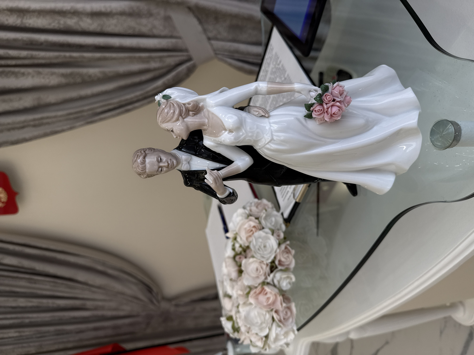 A porcelain figurine of a bride and groom stands on a glass-topped table. The groom, on the left, wears a black tuxedo with a white shirt and bowtie. The bride, on the right, wears a white wedding gown with long sleeves and a veil. She holds a bouquet of pink roses. To the left of the figurine is a large arrangement of white and pink roses. Several documents and a pen are visible on the table behind the figurines. In the background, gray curtains are drawn. The setting appears to be indoors.